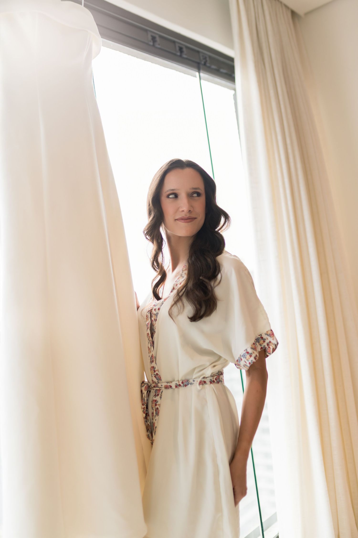 Bride in white robe with floral trim standing by window with curtains on wedding morning.