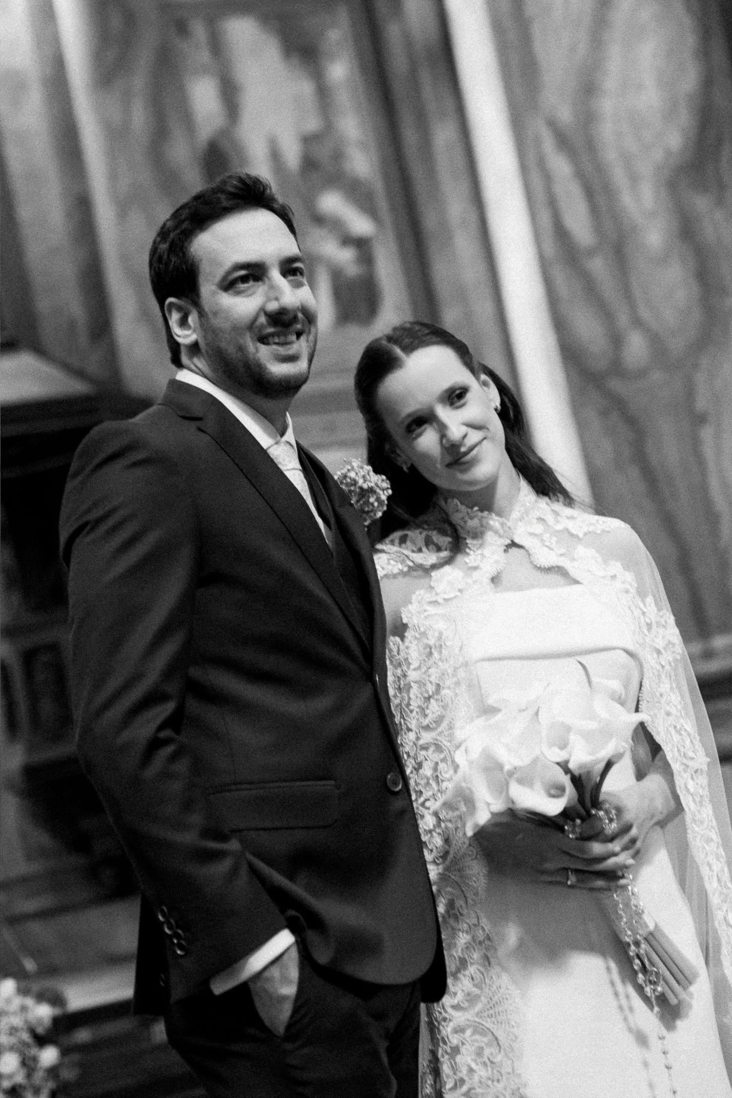 Black and white wedding photo of smiling bride holding bouquet and groom in suit posing together at elegant venue.