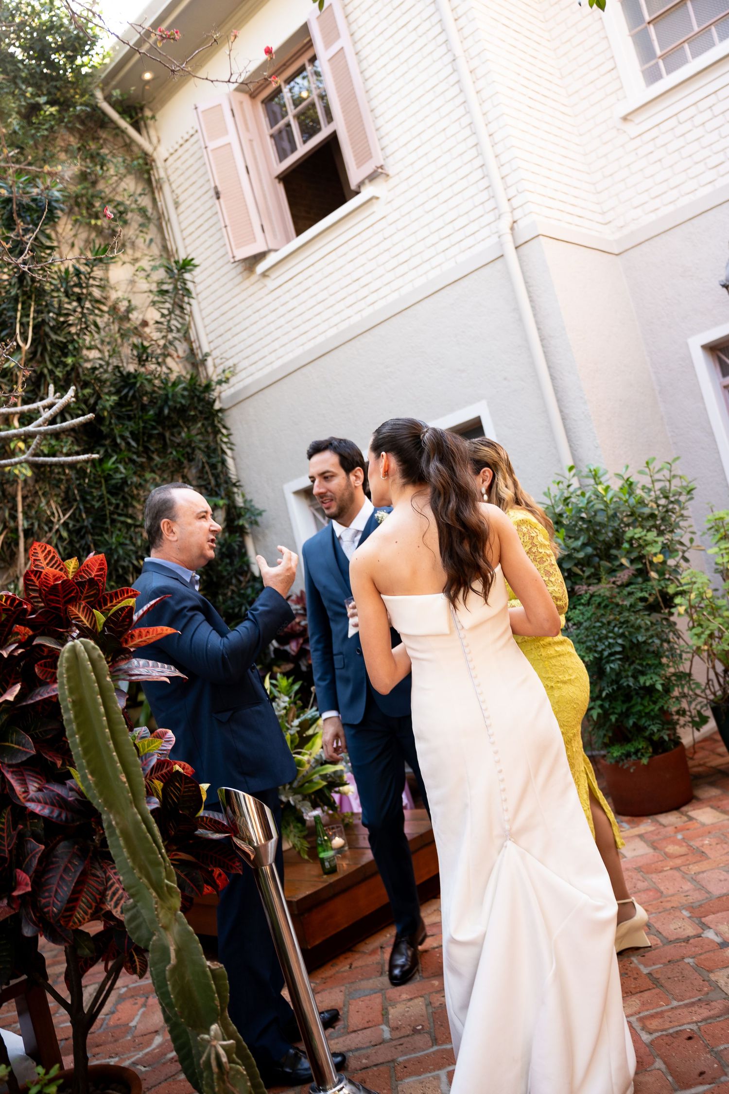 Bride in white gown and groom in blue suit greet guests in a charming outdoor courtyard setting.
