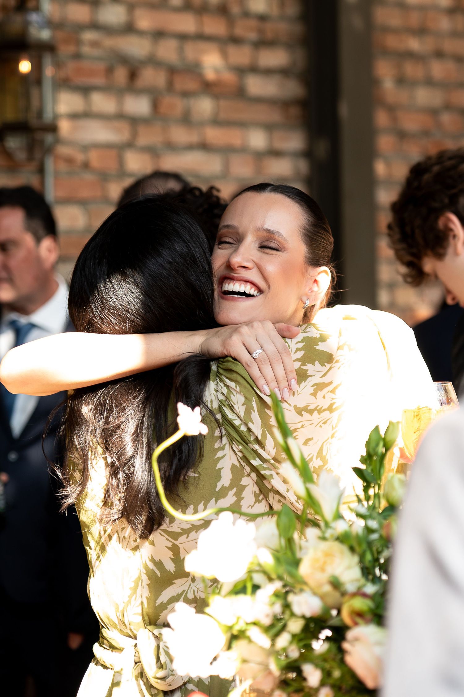 Two women share a joyful hug at a wedding, one laughing warmly while holding a bouquet of white flowers.