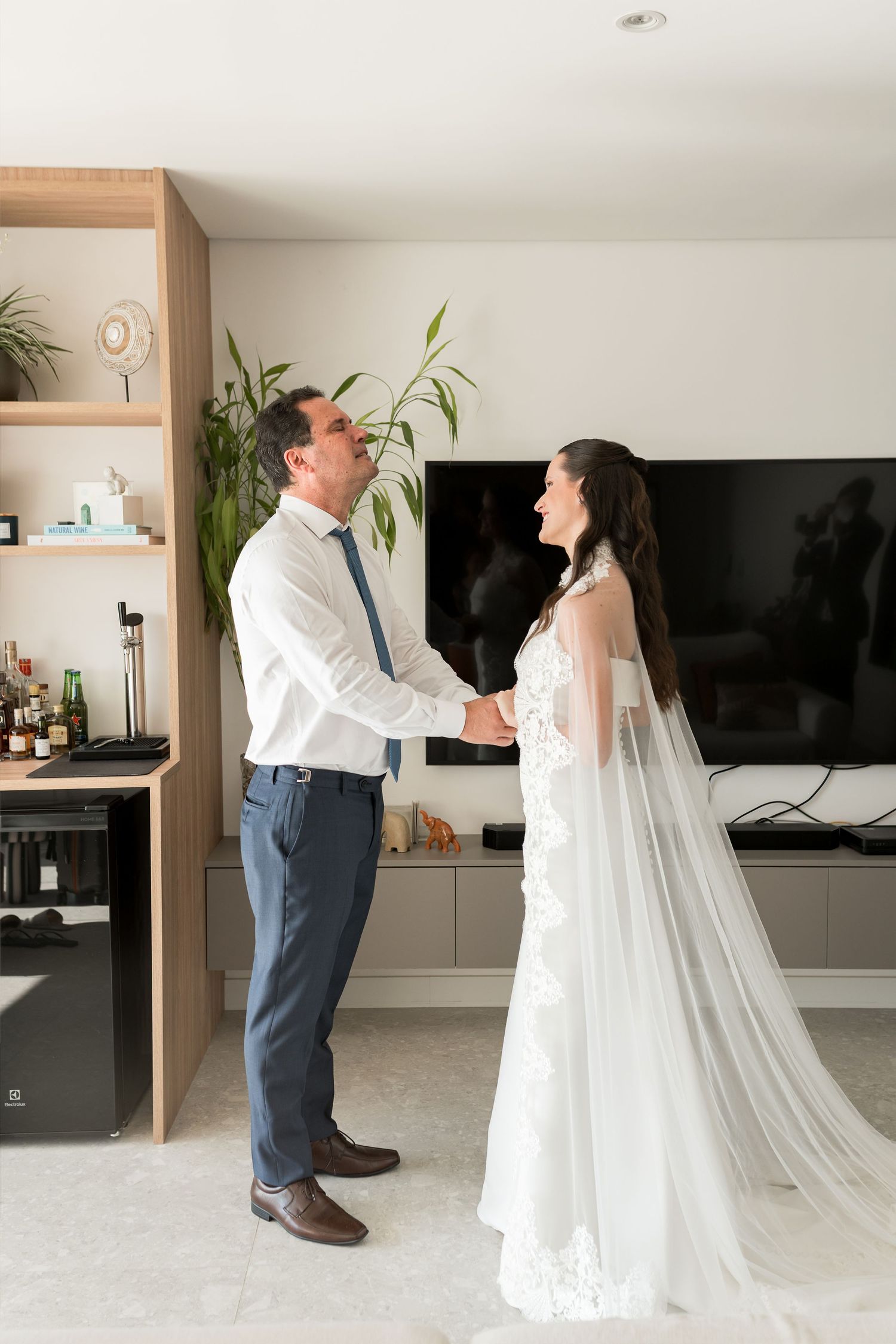 Bride and groom share a joyful moment holding hands in a modern living room during wedding day preparations.