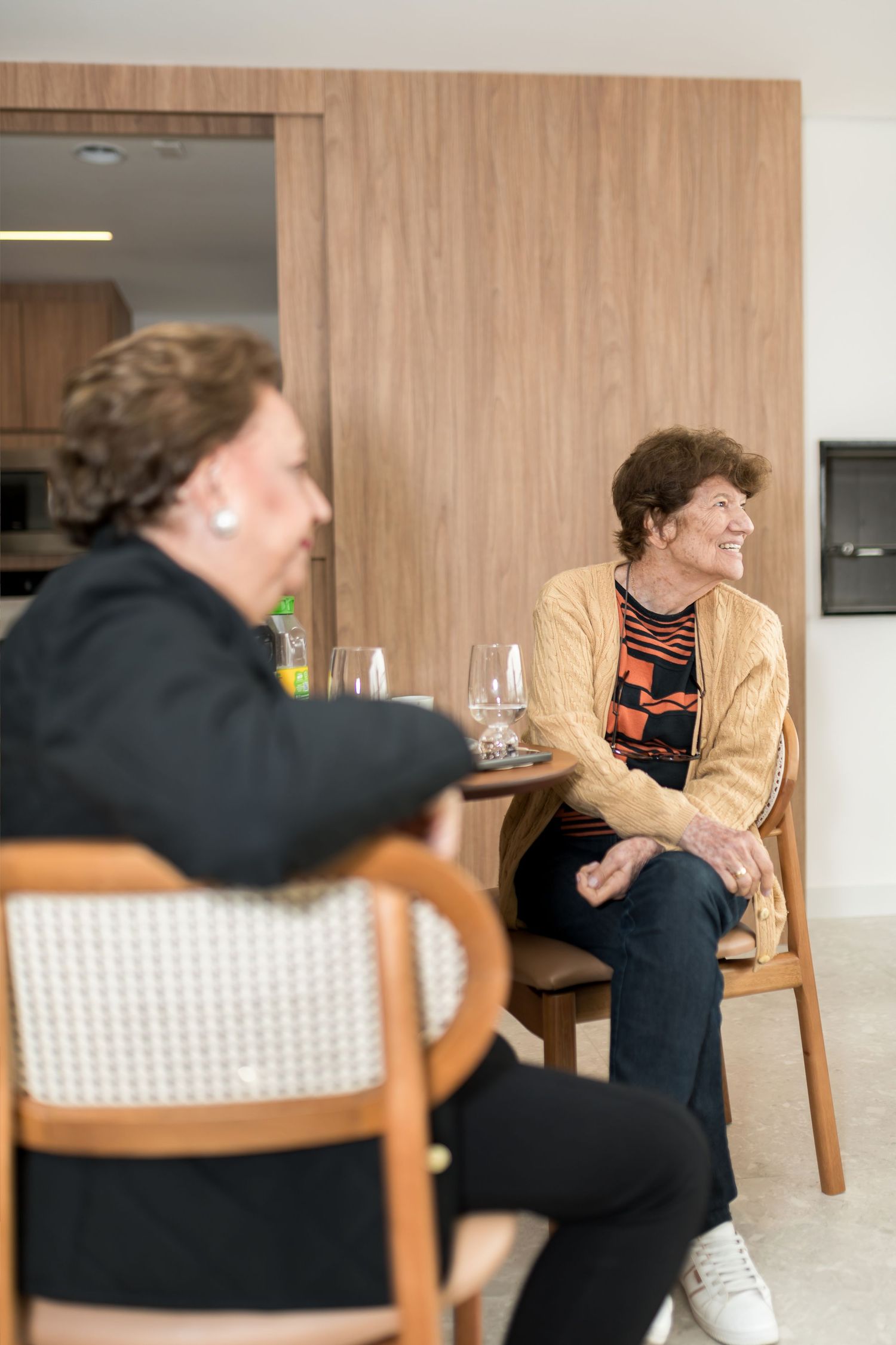 Two elderly women sitting on wooden chairs, smiling and conversing in a modern living space with warm wood paneling.