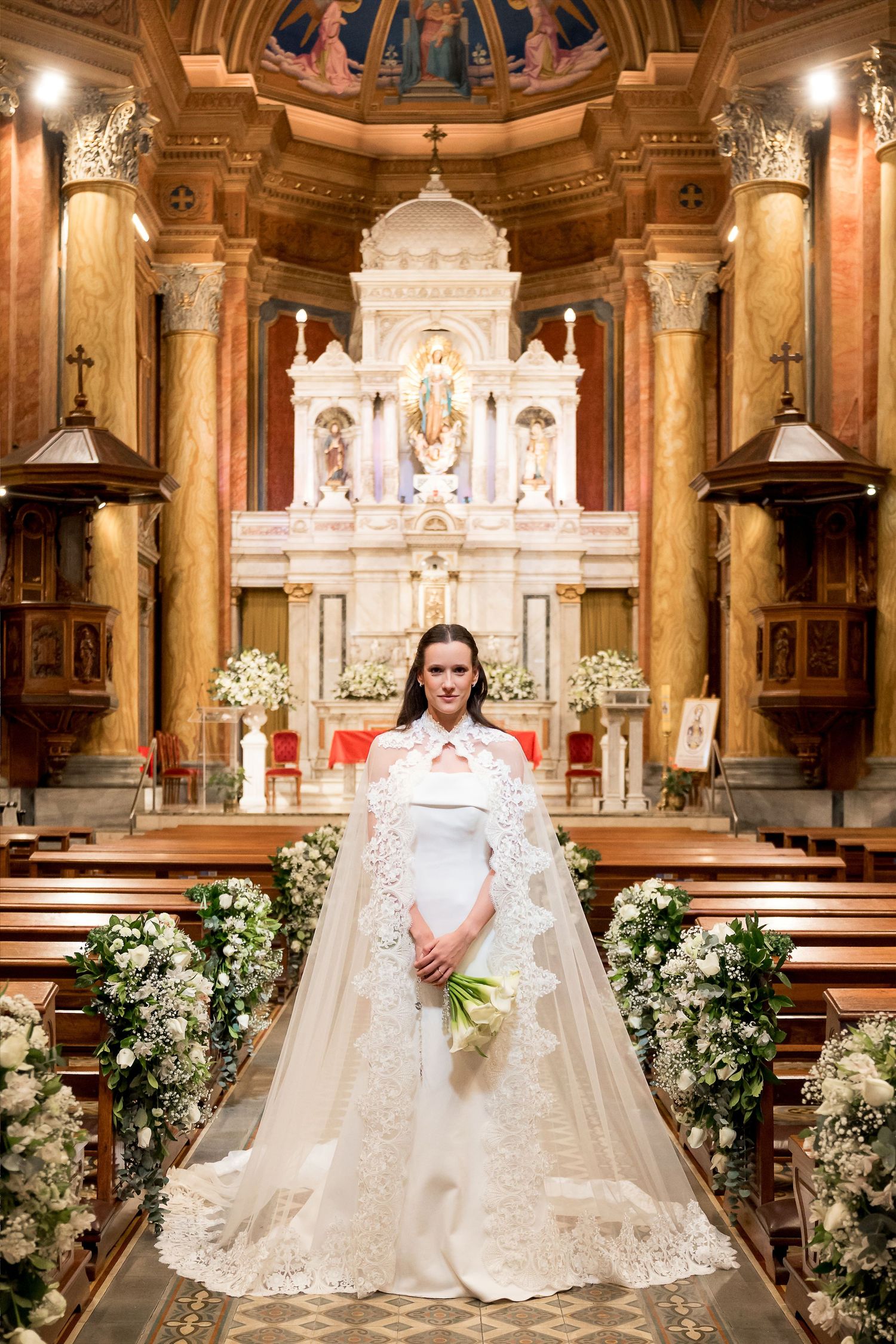 Elegant bride in white lace cape gown holding bouquet at ornate church altar with floral decorations.