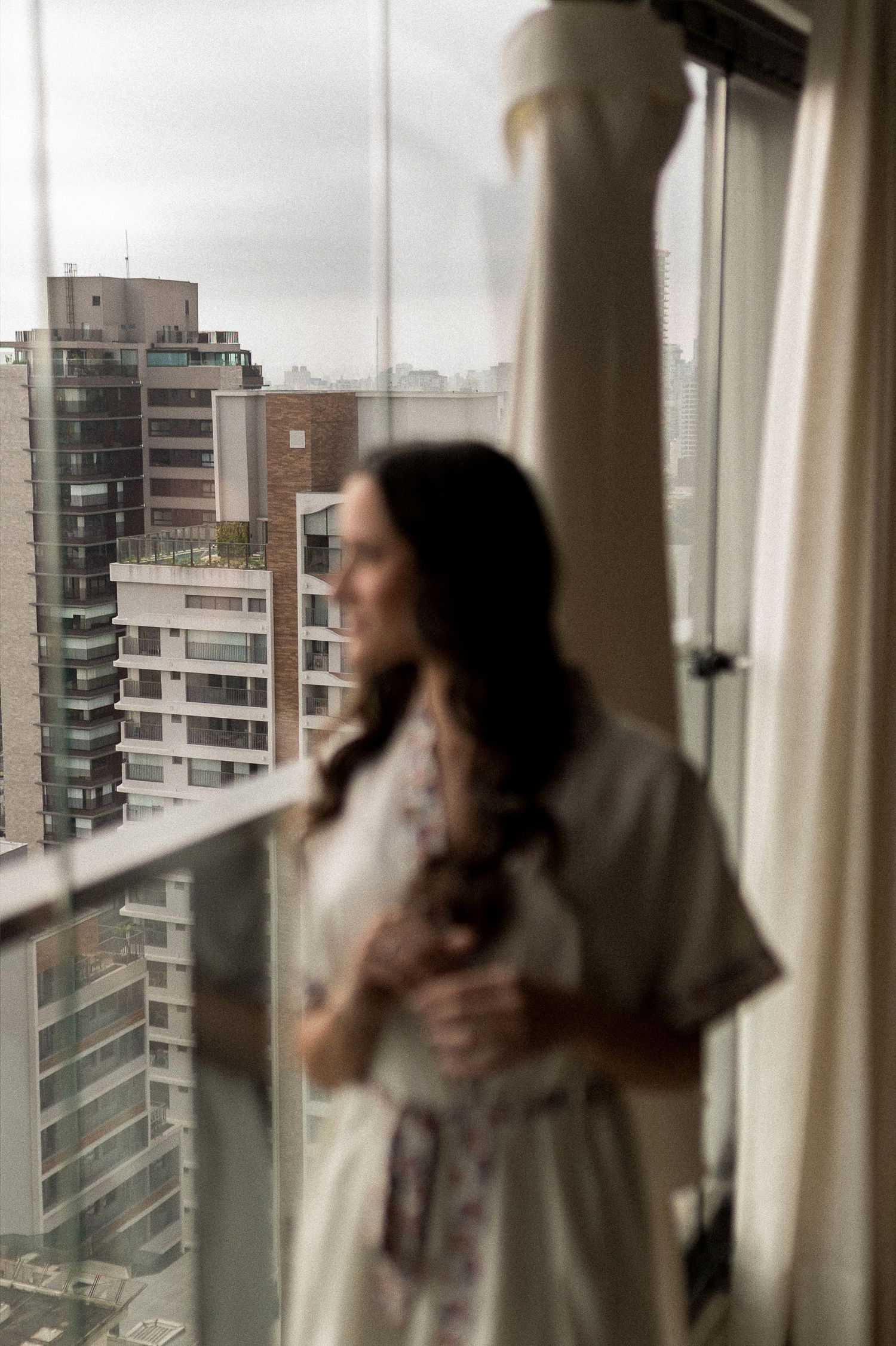 Bride in robe gazing out hotel window with wedding dress hanging nearby, city skyline in background.