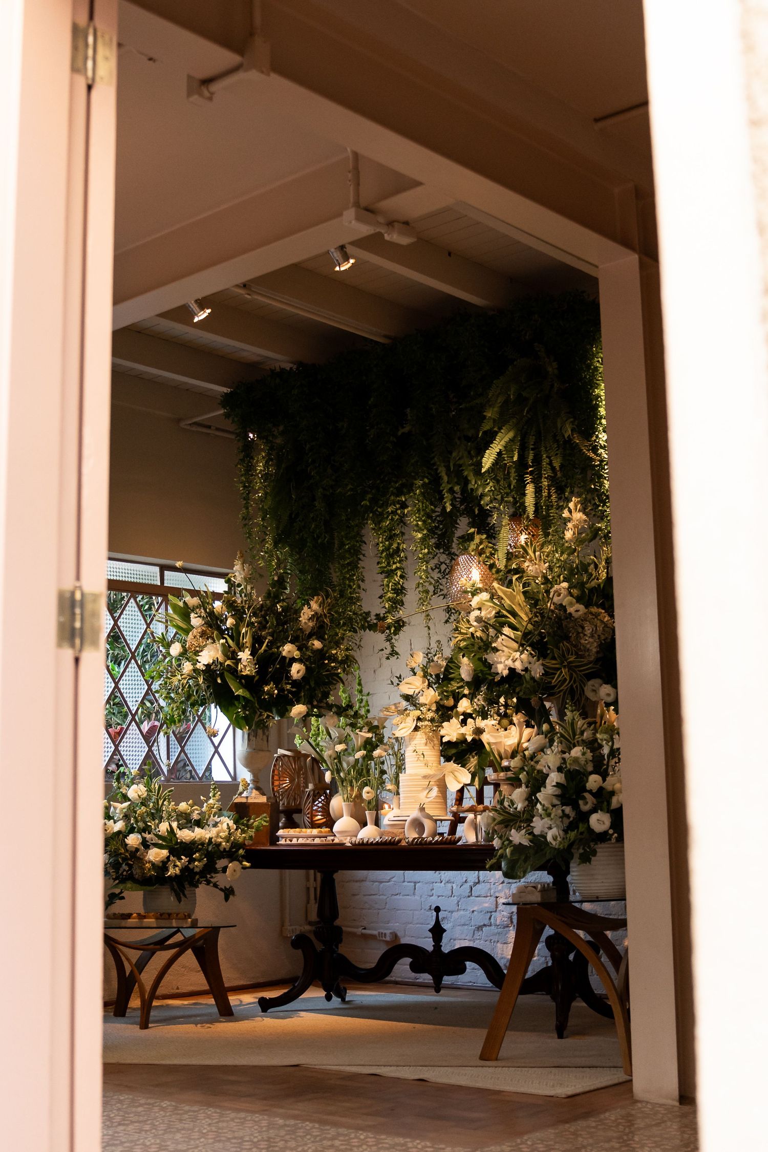 Elegant wedding dessert table adorned with lush greenery, white florals, and hanging vines in a warmly lit venue.