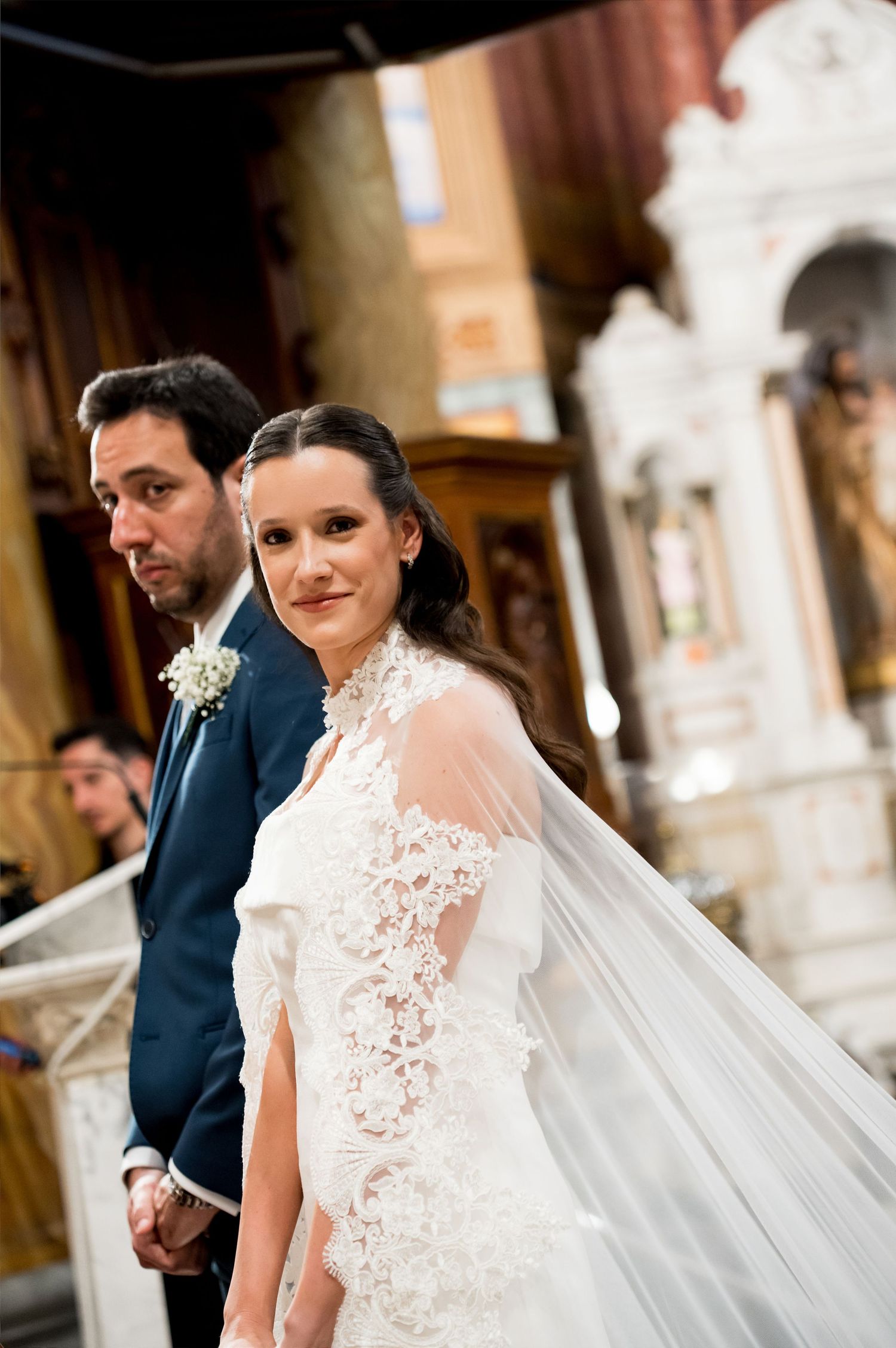 Bride in lace wedding gown and veil smiles at camera during church ceremony with groom in blue suit behind her.