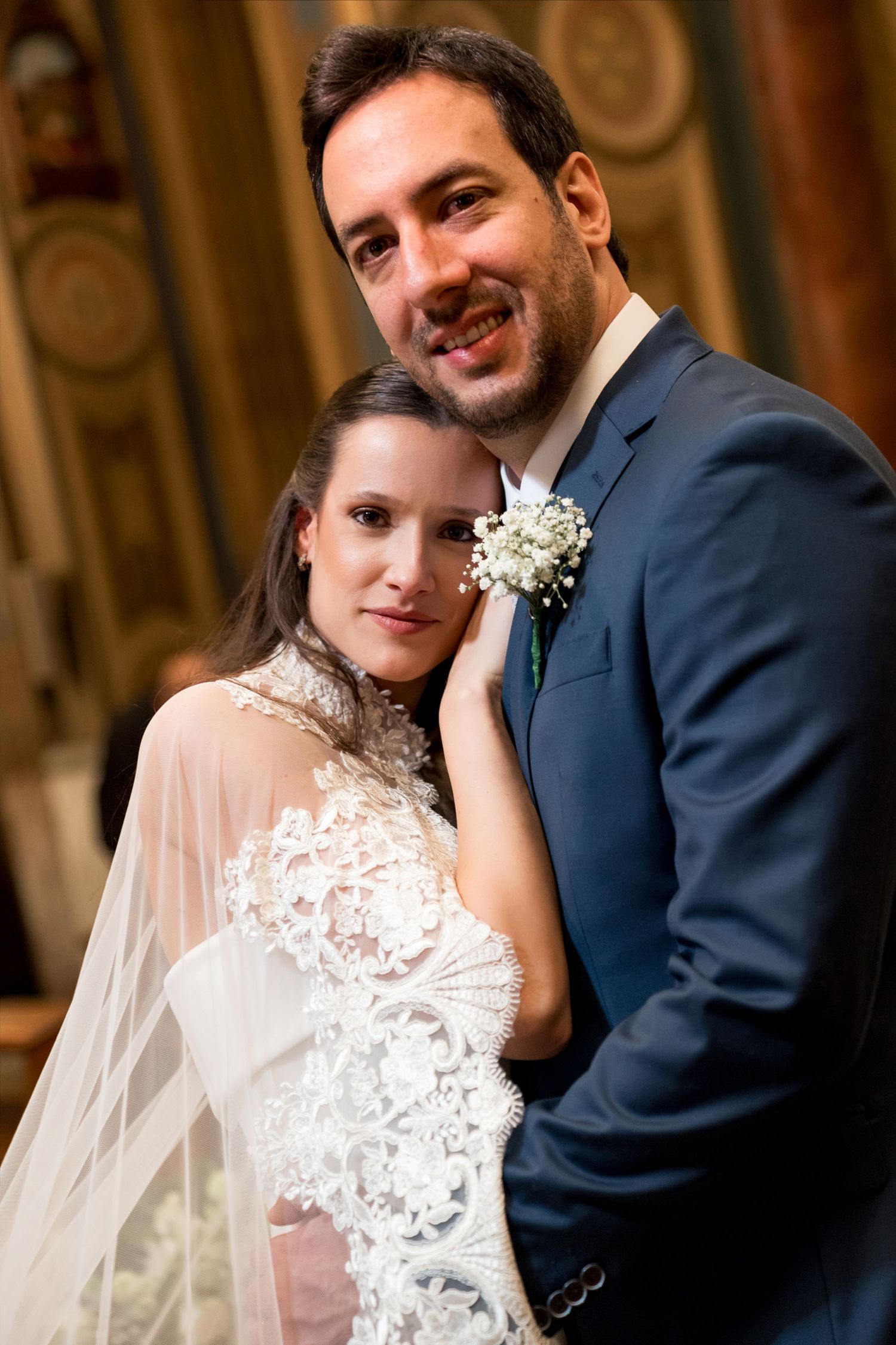 Bride in lace gown and groom in navy suit pose together at elegant wedding ceremony venue.