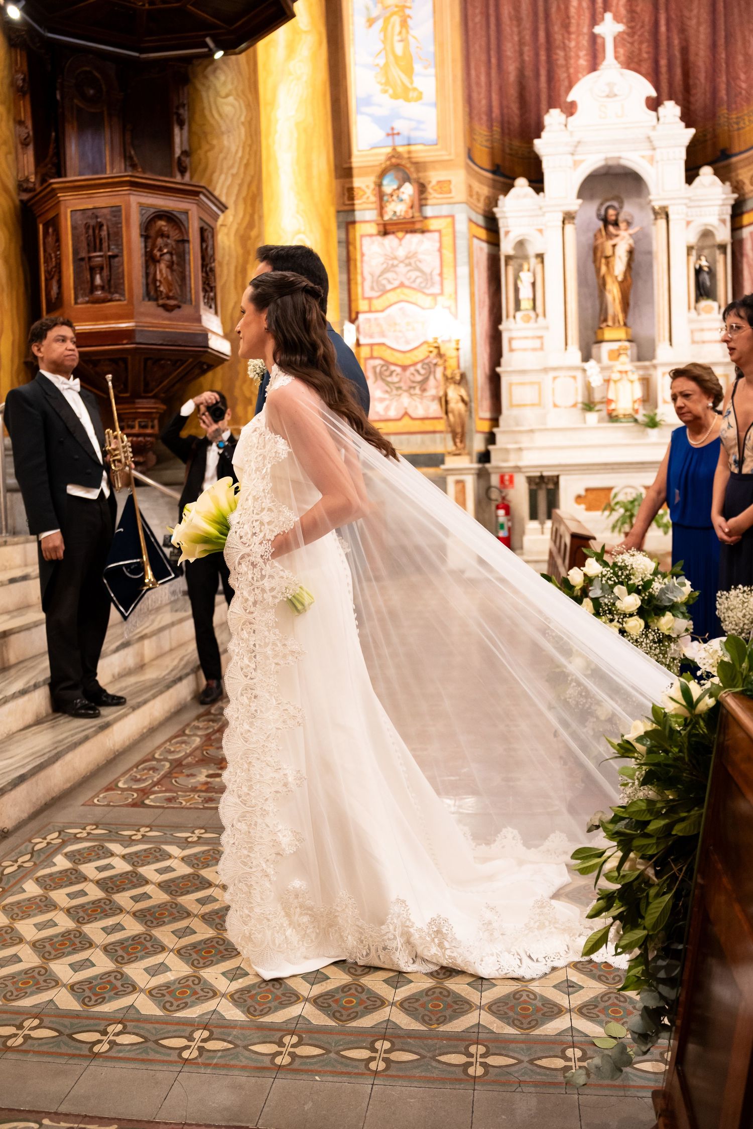 Bride in lace wedding gown with flowing veil stands at church altar holding bouquet during ceremony.