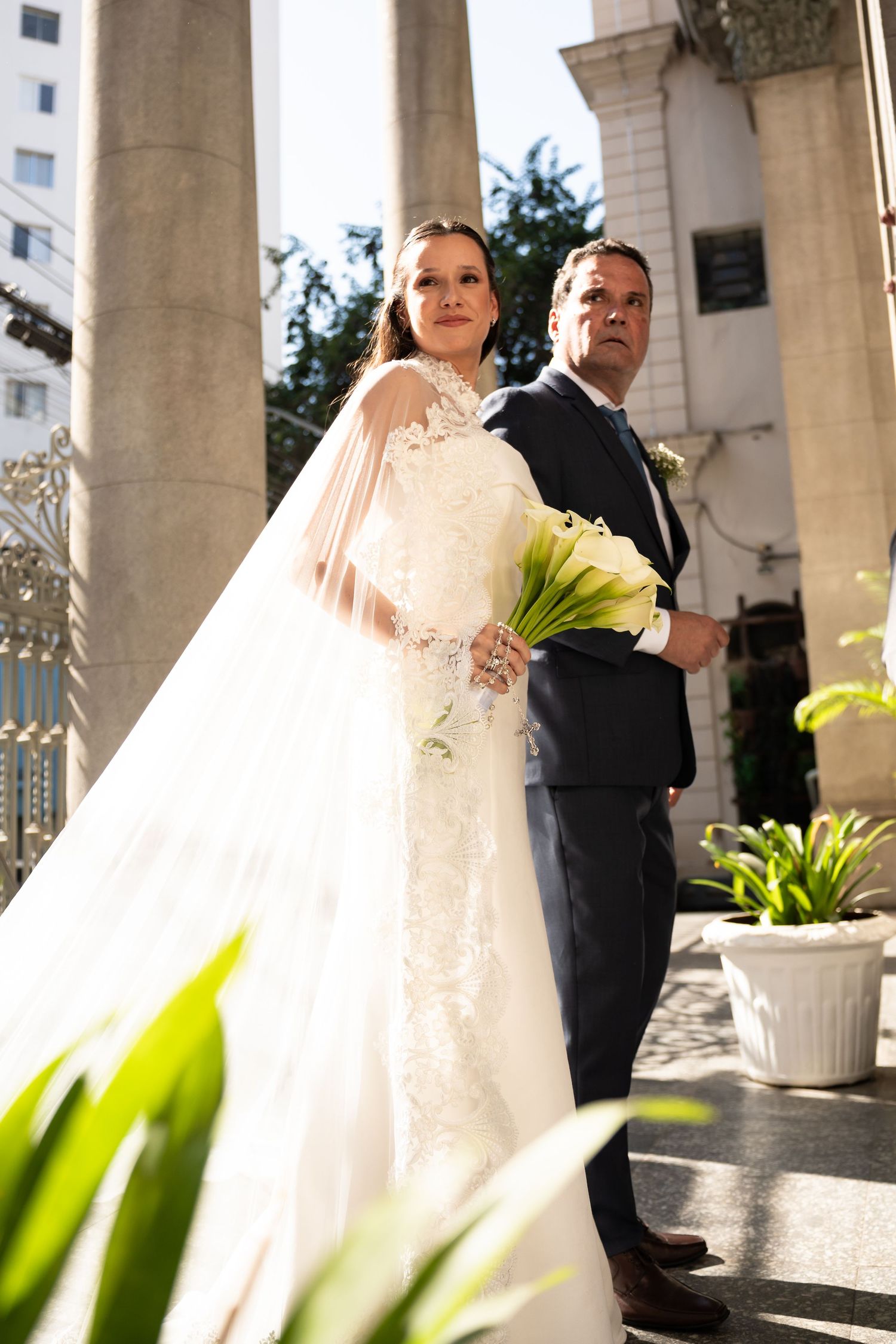 Bride in lace gown with veil holds white calla lily bouquet beside suited groom at elegant venue.