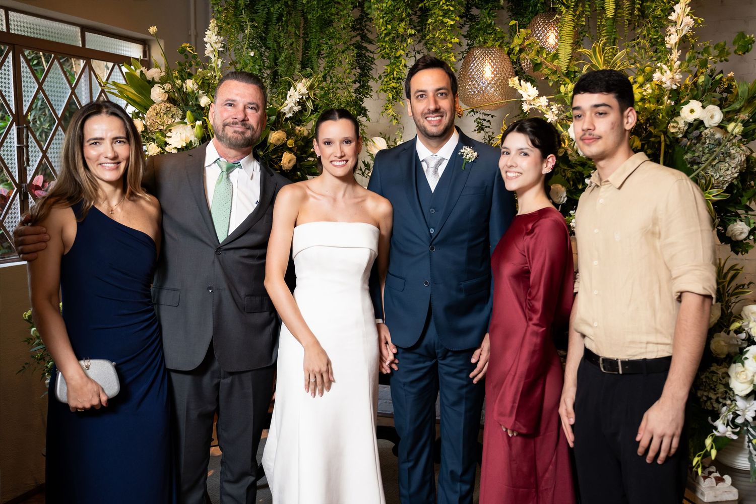 Wedding couple poses with family members in formal attire before floral backdrop at elegant reception venue.