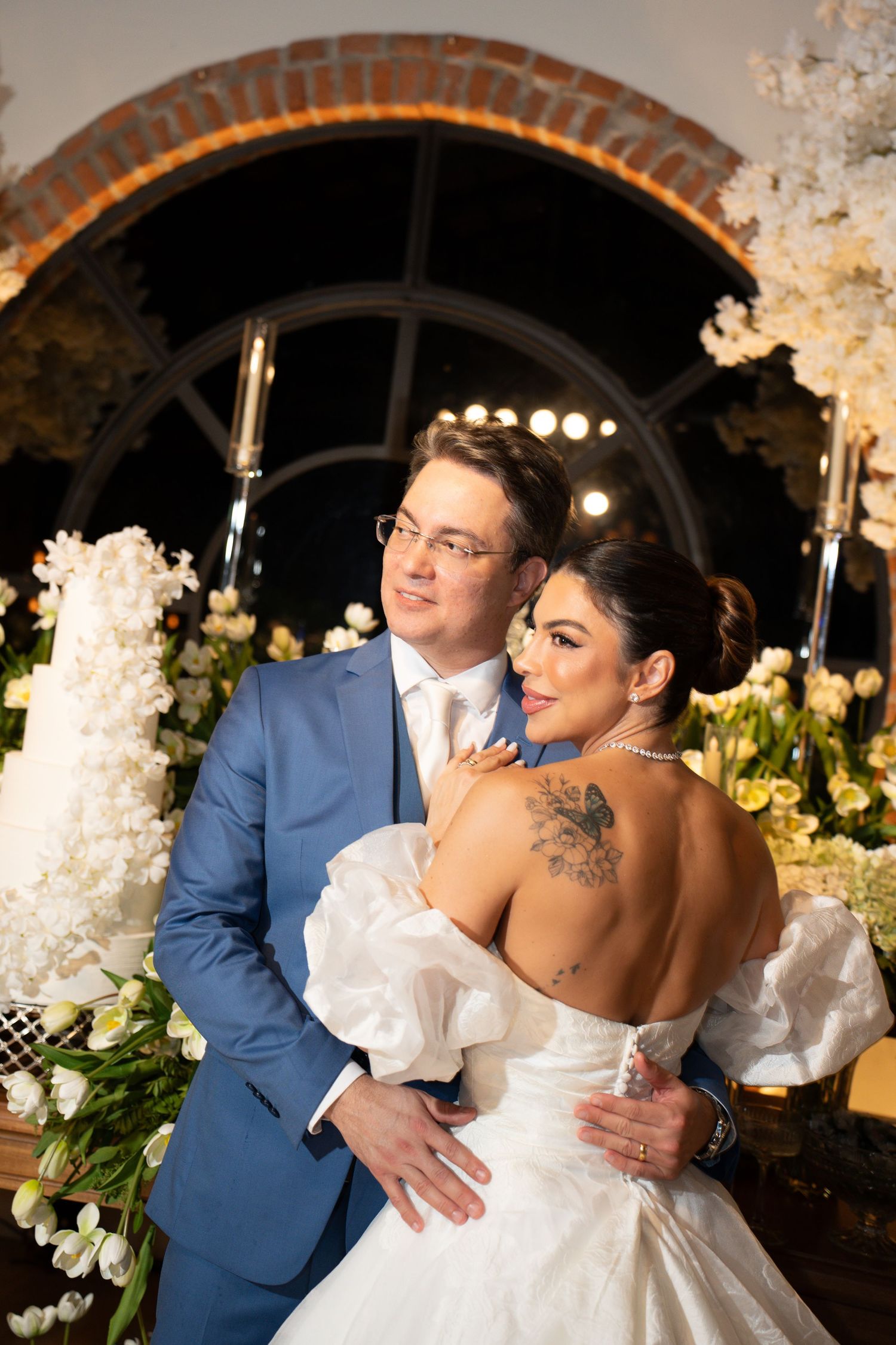 Bride in white off-shoulder gown with floral tattoo embraces groom in blue suit before elegant floral wedding backdrop.