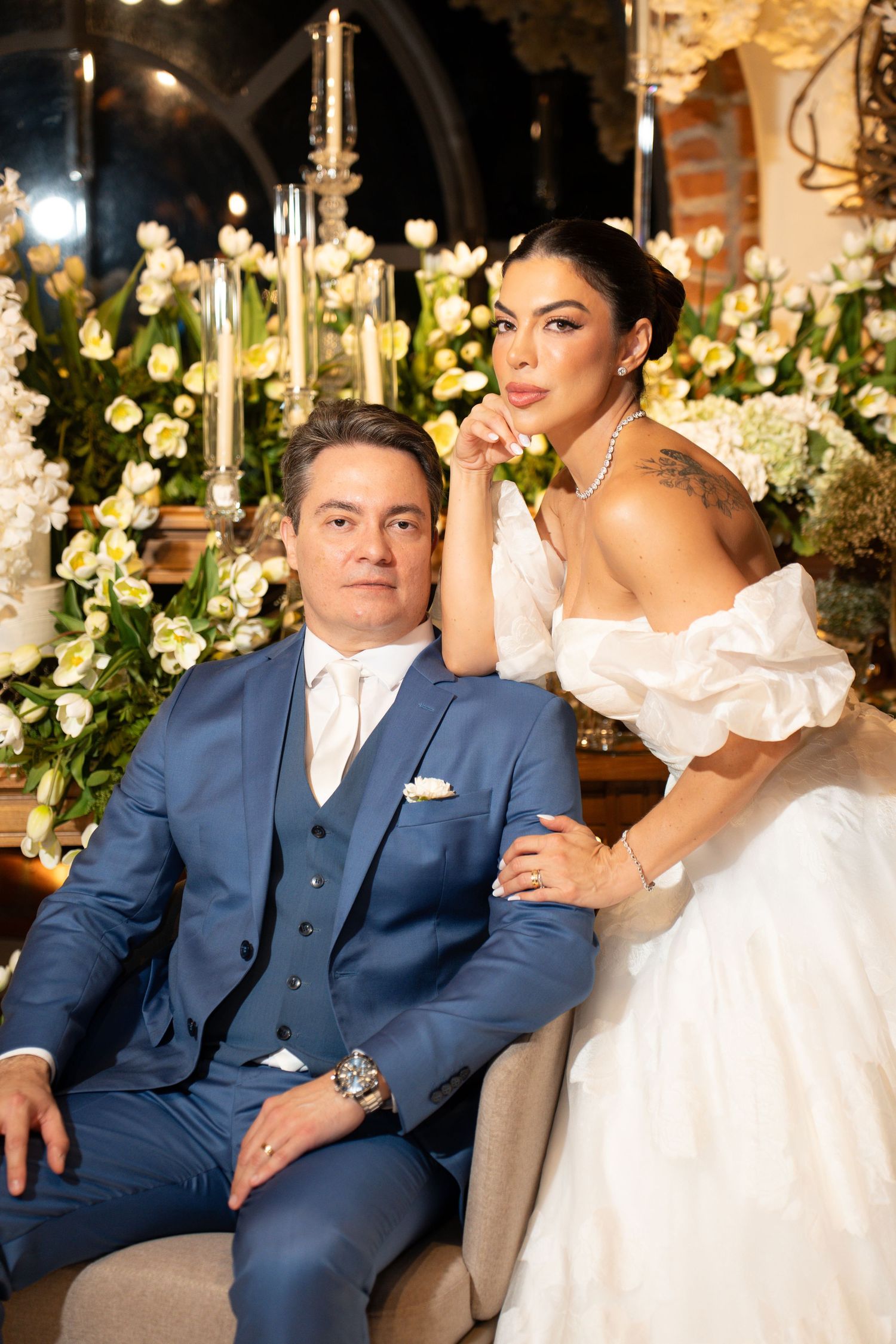 Elegant bride and groom posing at wedding reception with white floral arrangements and candlelight backdrop.
