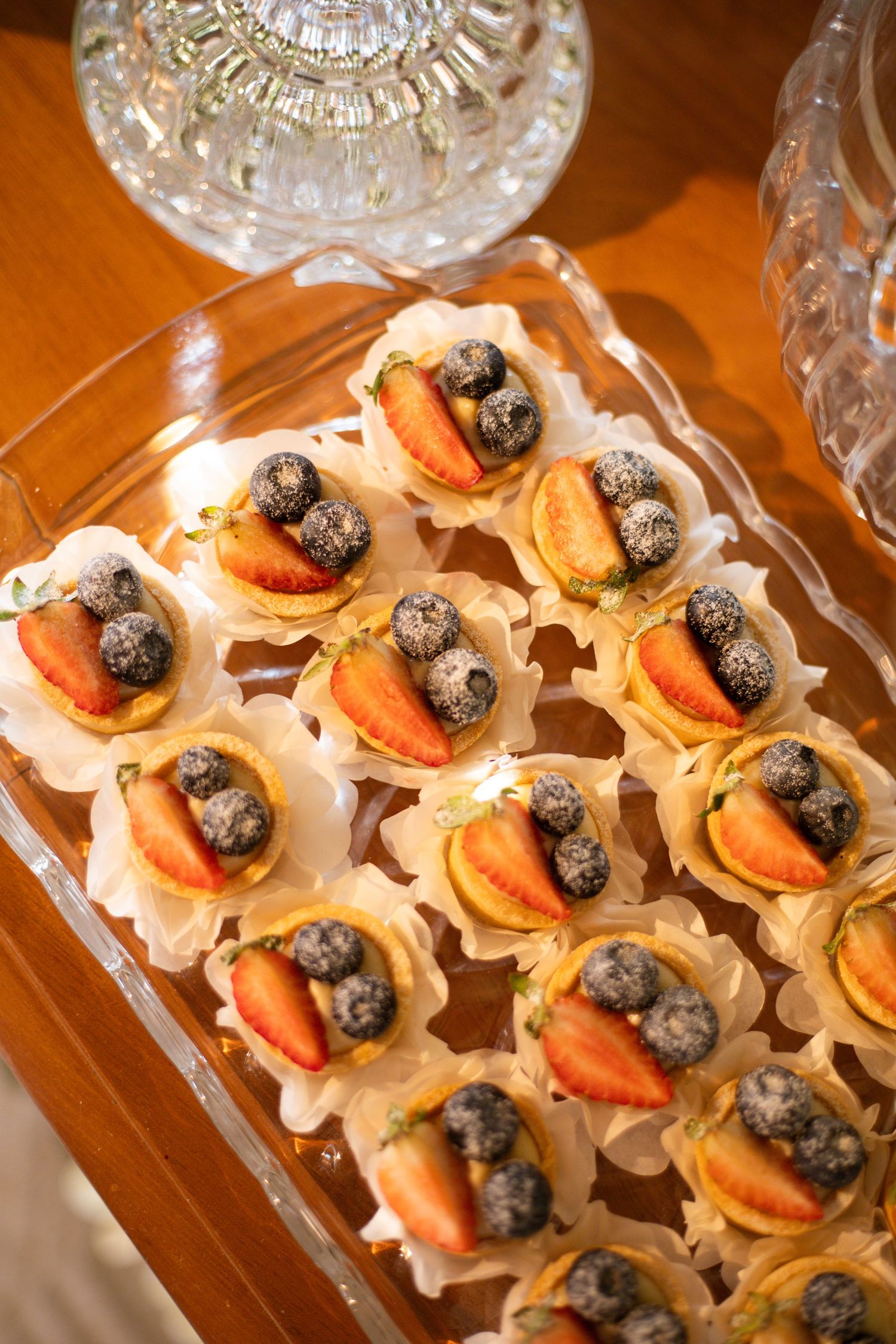 Elegant glass tray of mini fruit tarts topped with fresh strawberries and blueberries dusted with powdered sugar.