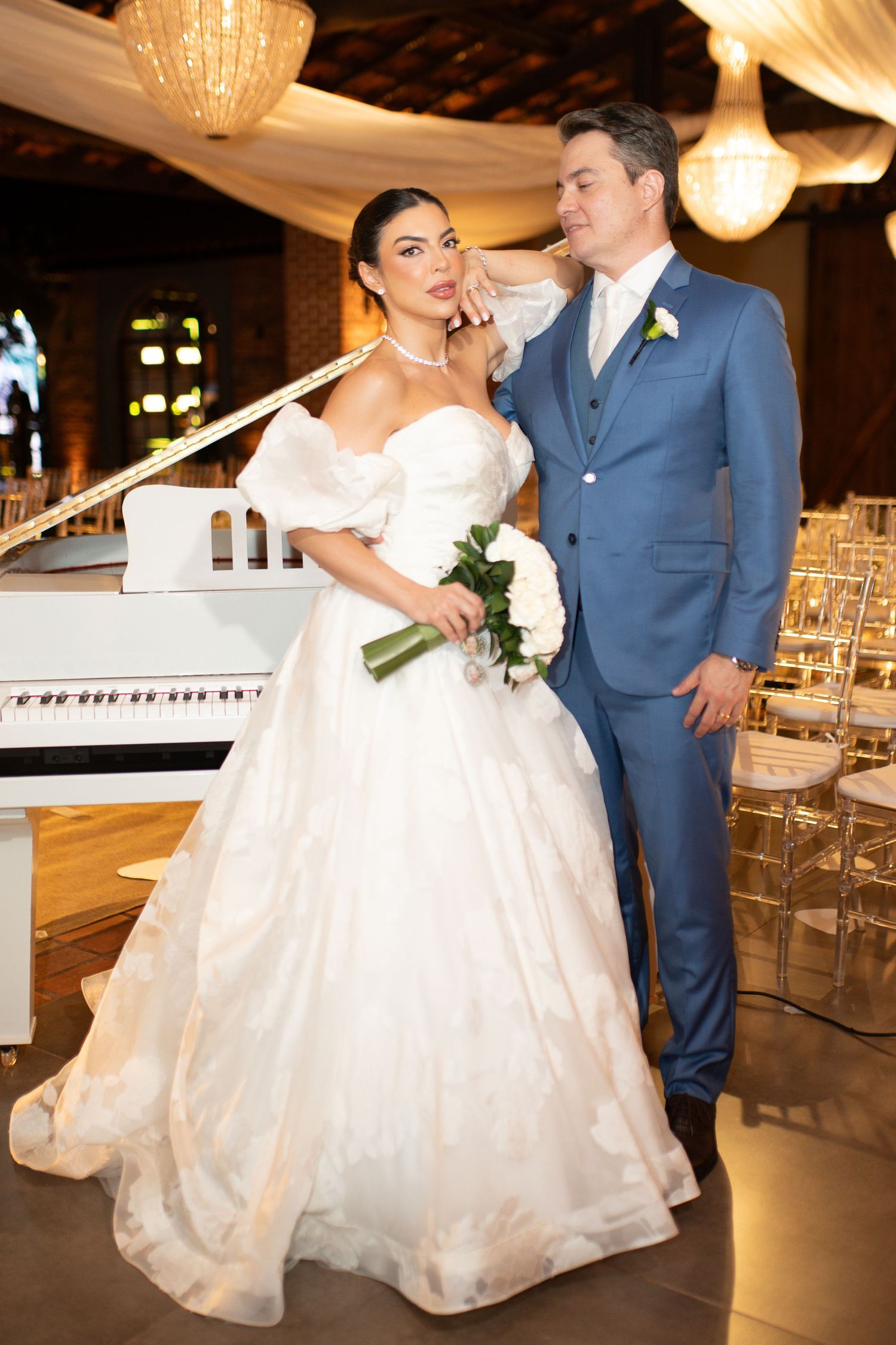 Bride in white ballgown holding bouquet stands with groom in blue suit near white piano at elegant wedding reception.