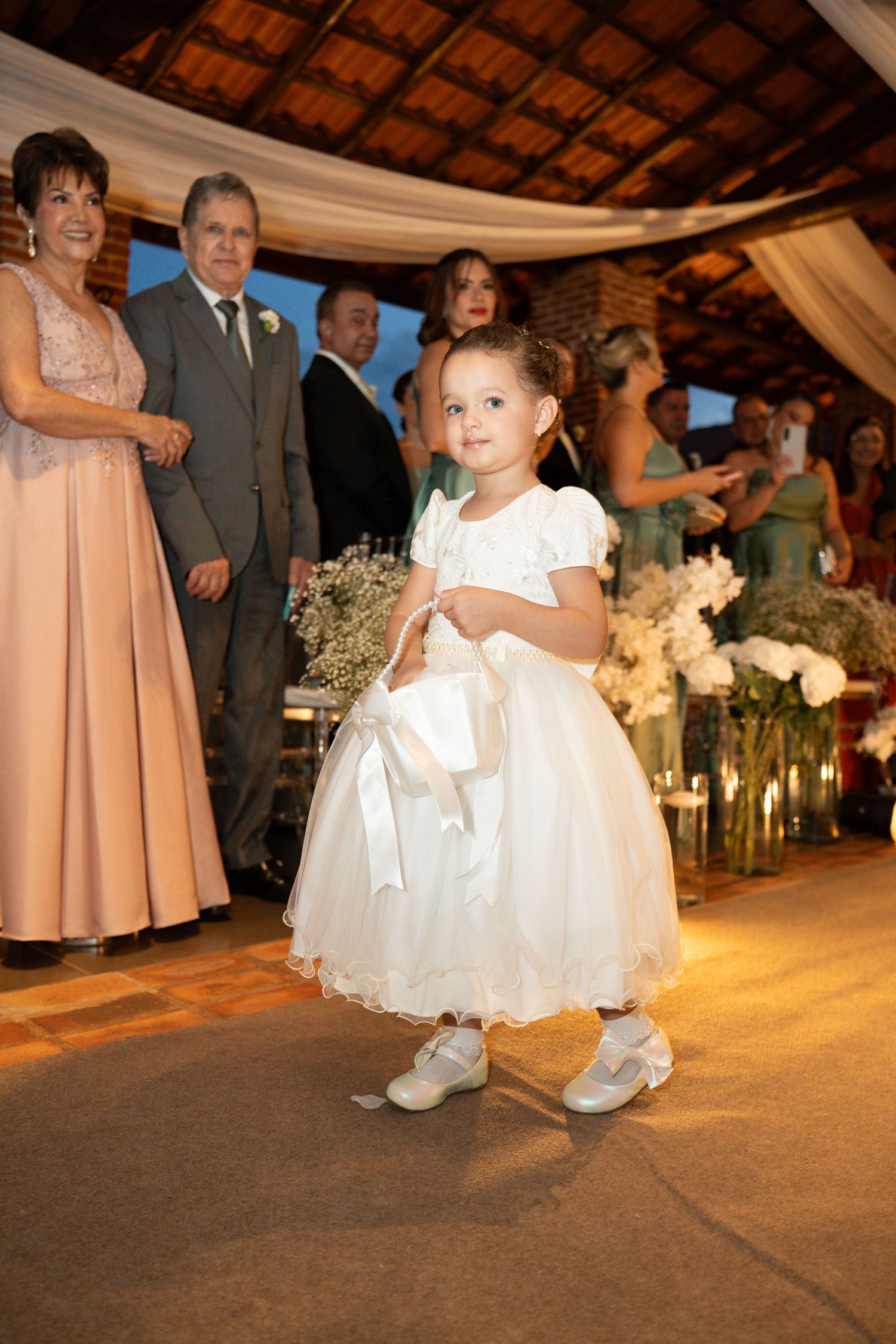 Young flower girl in white dress walking down wedding aisle holding bouquet, guests watching in background.