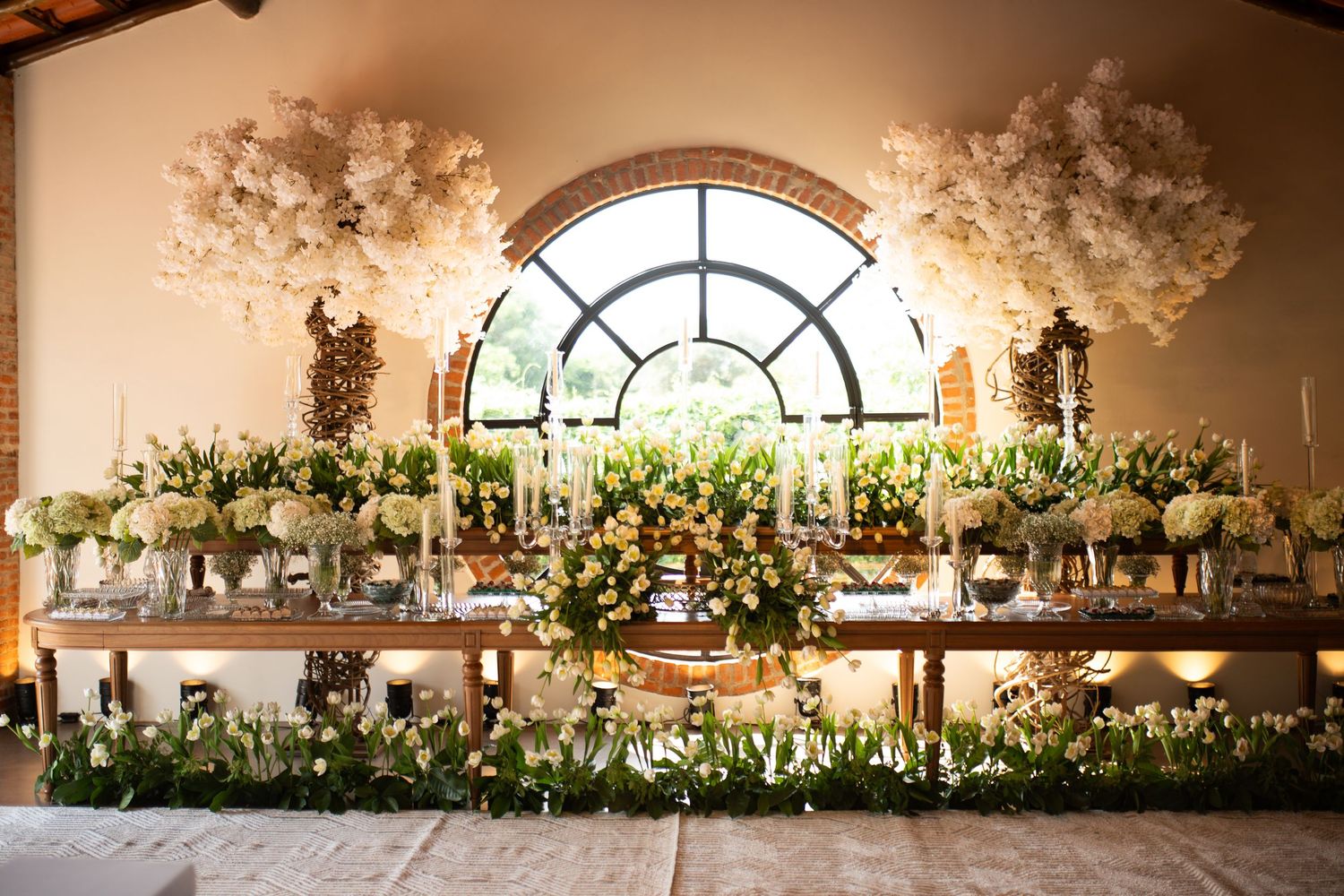 Elegant wedding reception table adorned with lush white floral arrangements, candles, and greenery beneath an arched window.