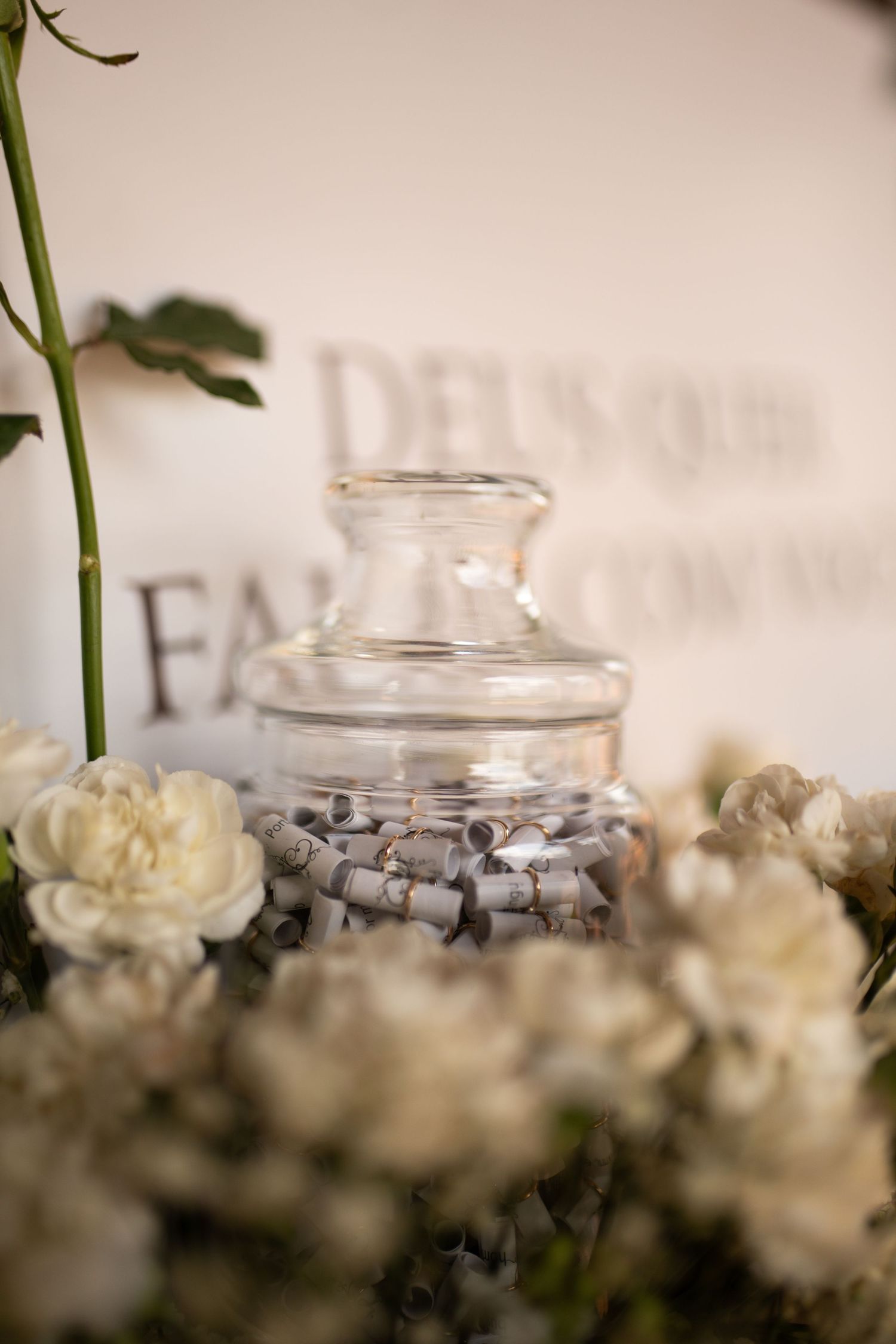 Glass jar filled with rolled paper notes surrounded by white peonies and roses at an elegant wedding reception.