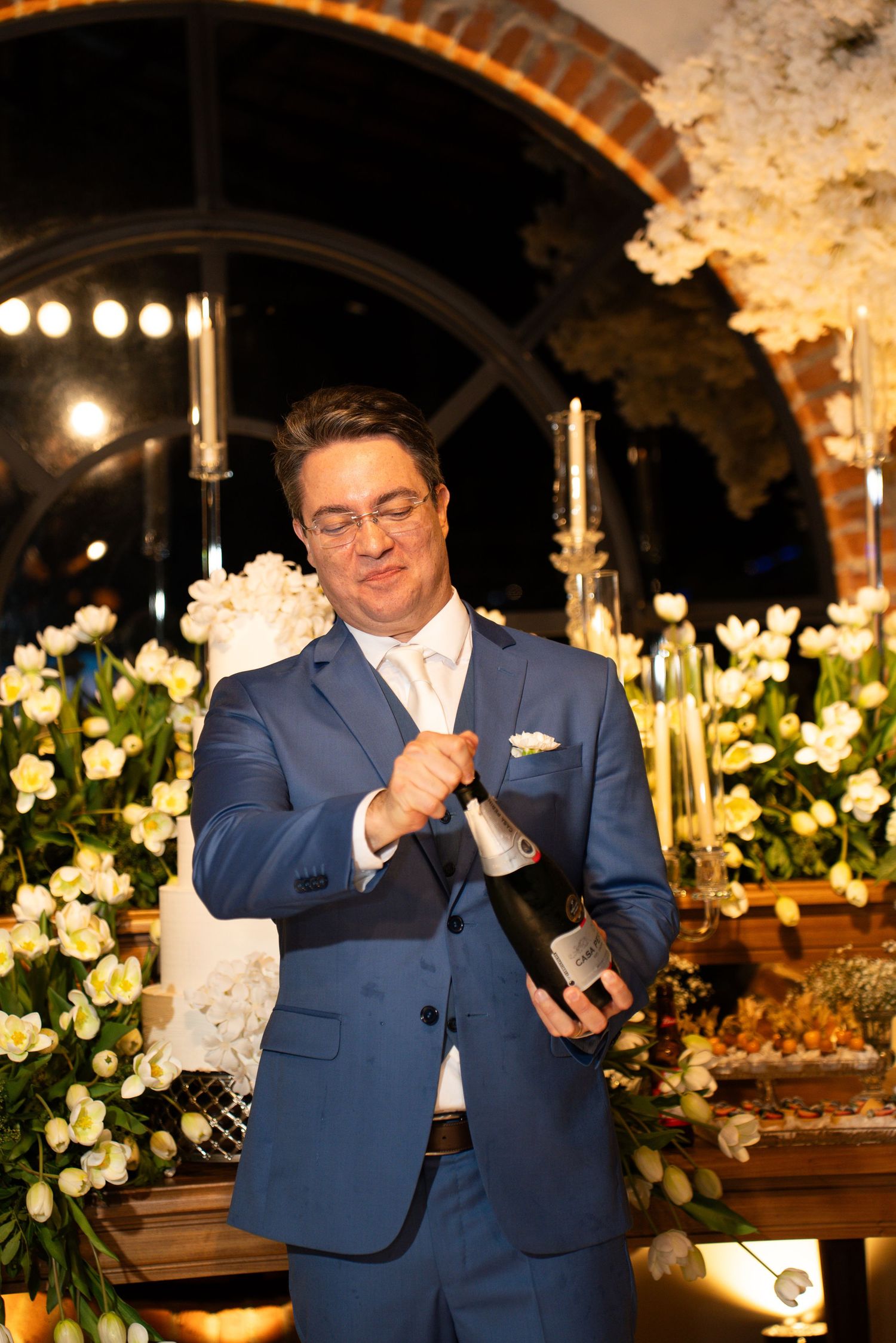 Man in blue suit opening champagne bottle surrounded by white floral arrangements at elegant event.