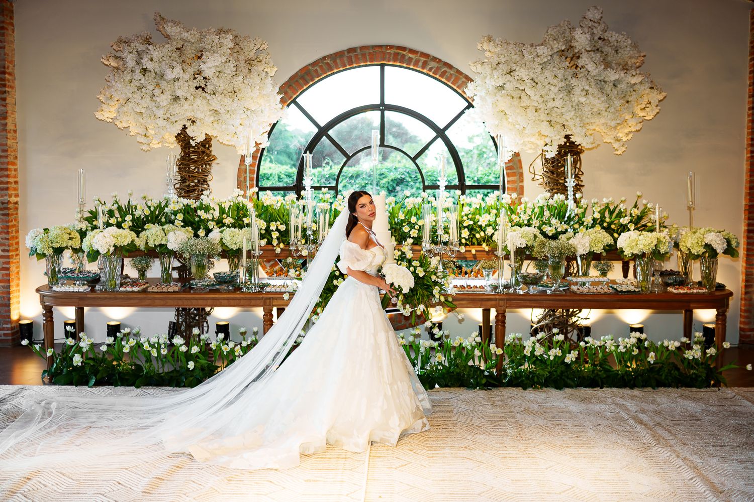 Bride in white gown stands before lush floral backdrop with greenery and white blooms at elegant wedding reception.