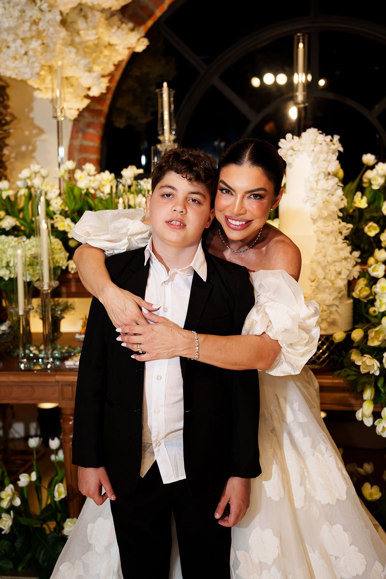A bride in a white off-shoulder gown hugs a young boy in a black suit at an elegant floral wedding reception.