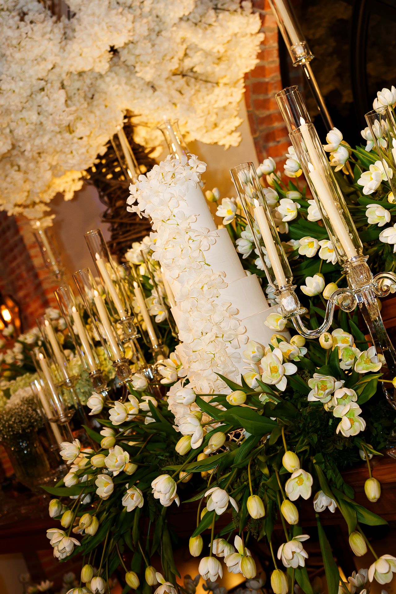 Elegant white wedding cake adorned with flowers, surrounded by white tulips and candelabras in a warmly lit venue.