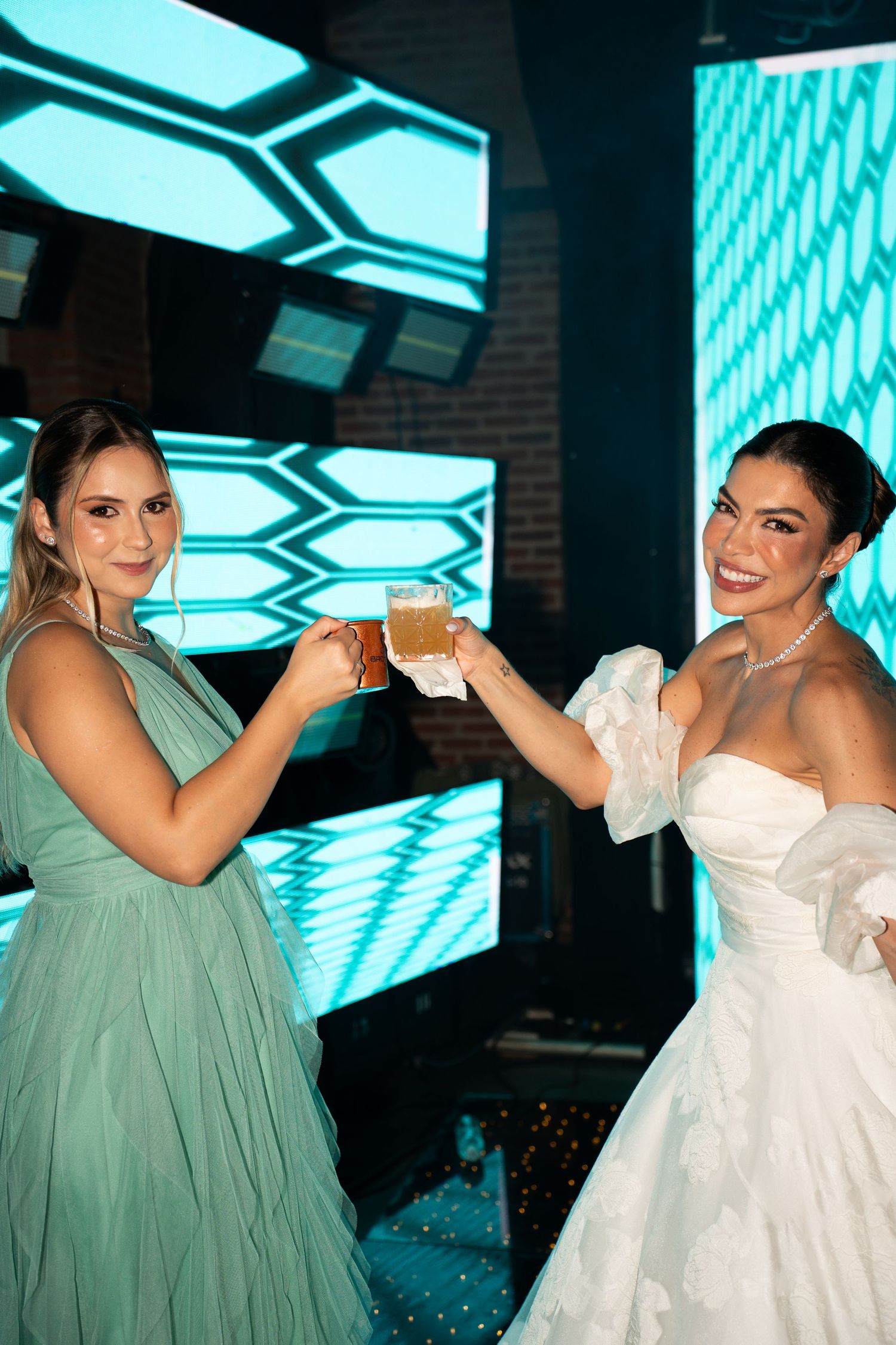 Bride in white gown and bridesmaid in teal dress toasting drinks at a wedding reception.
