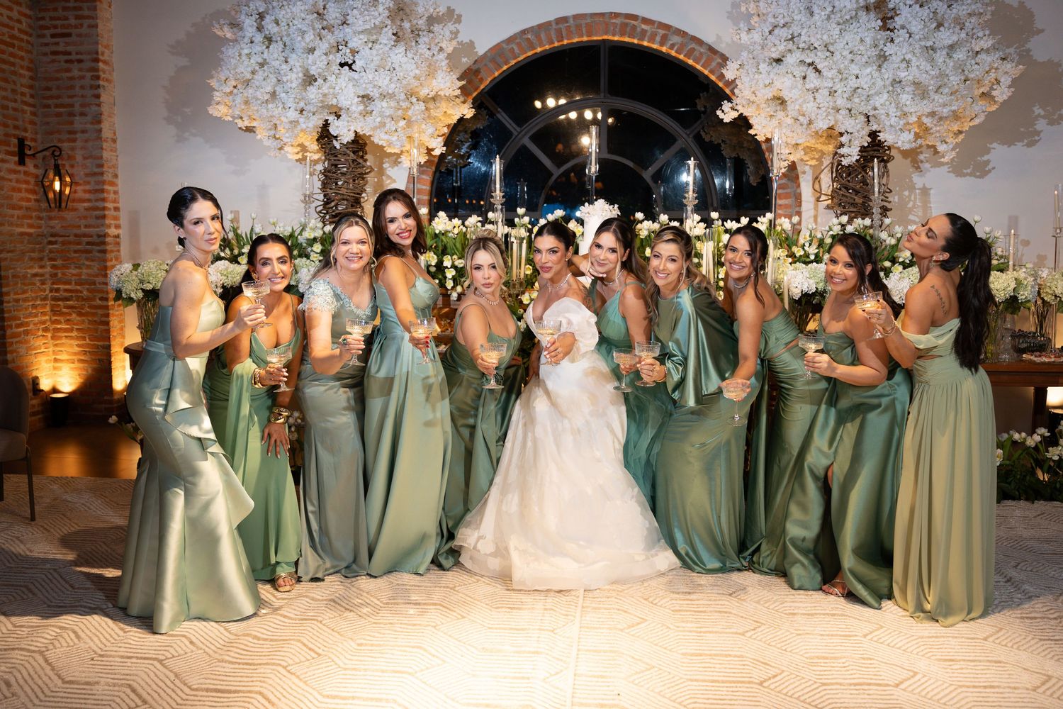 Bride in white gown poses with bridesmaids in sage green dresses at elegant floral wedding reception.