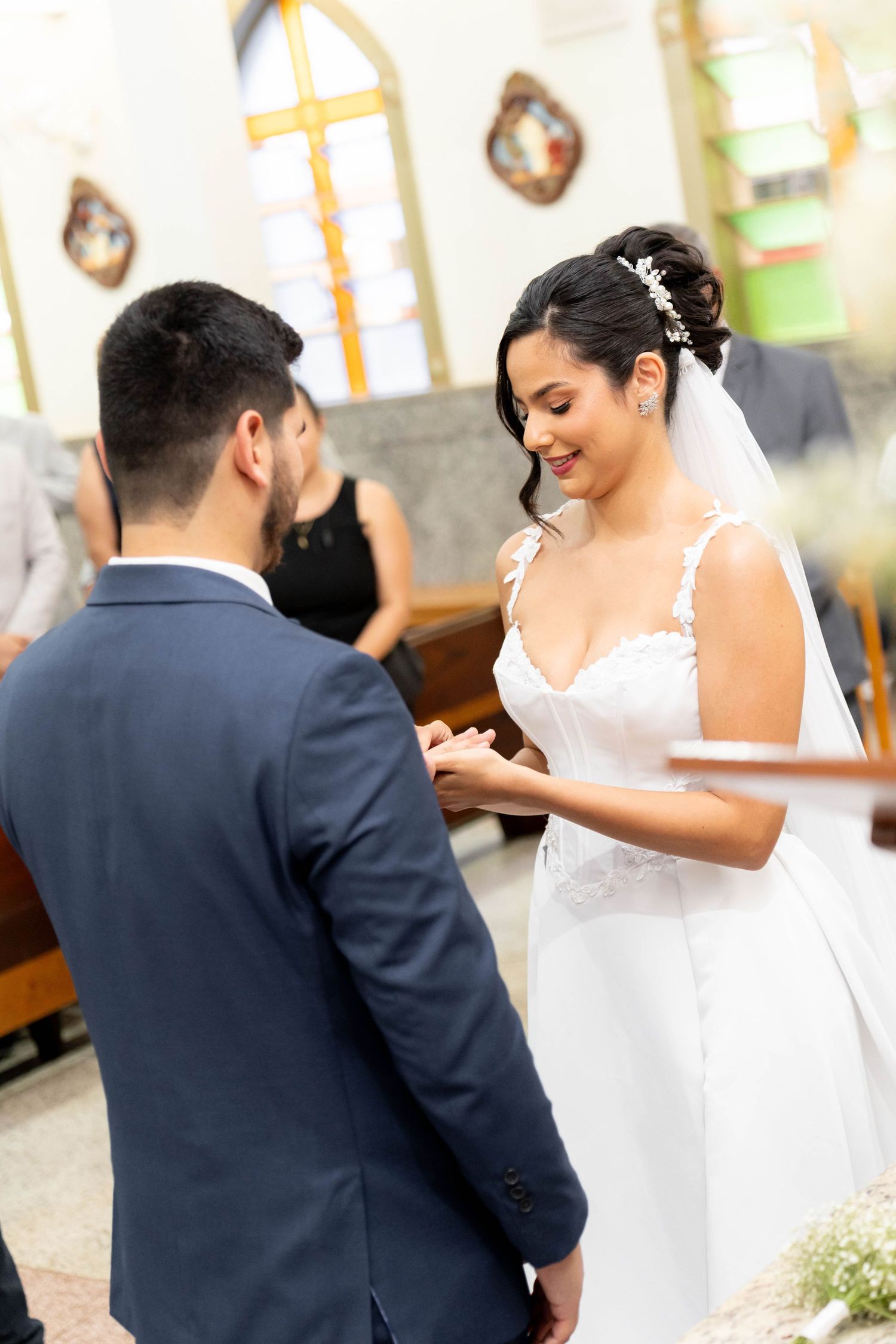 Bride smiling while placing wedding ring on groom's finger during church ceremony, wearing white lace gown and veil.