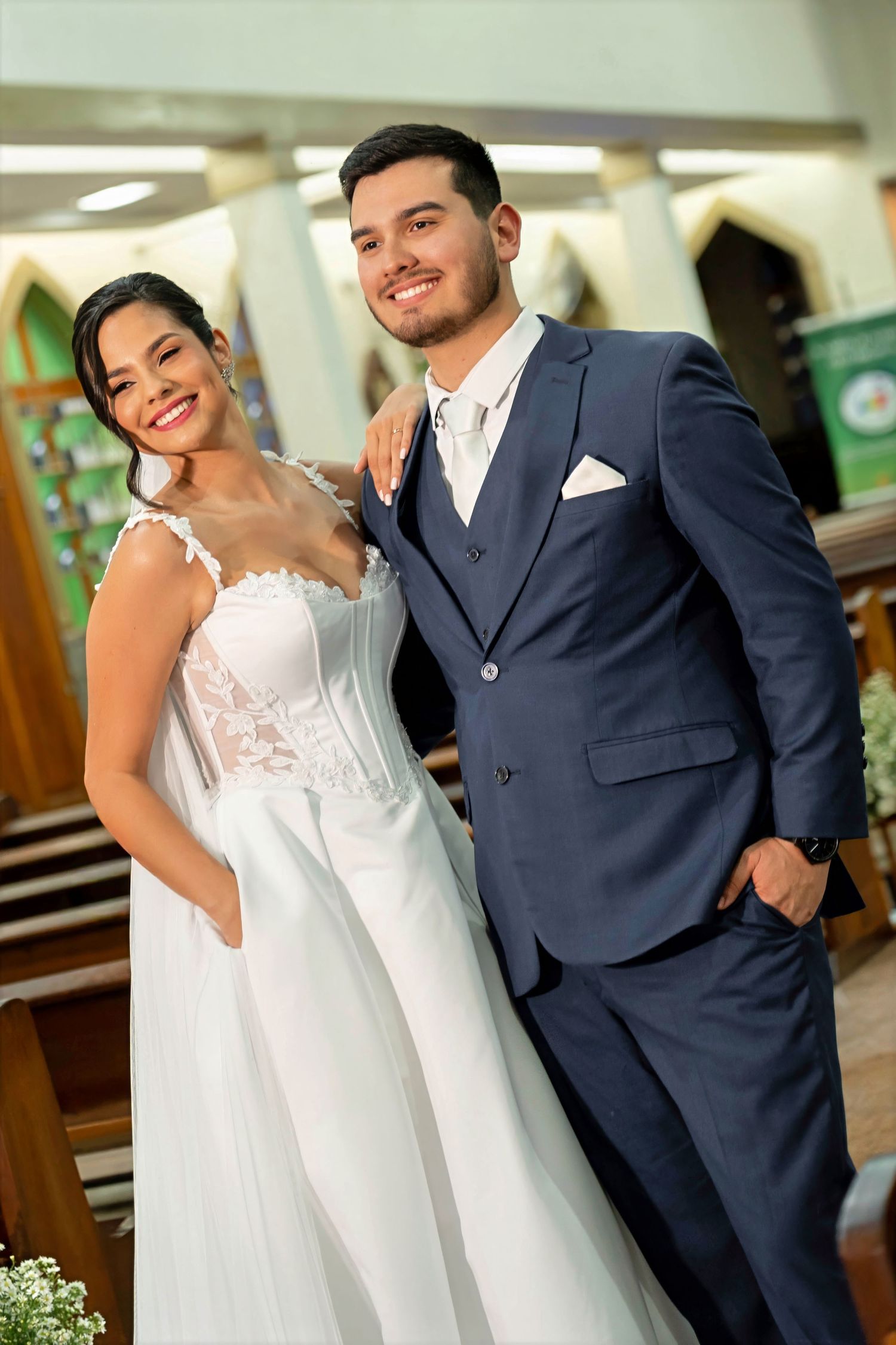 A smiling bride in a white lace gown and groom in a navy suit pose together inside a church on their wedding day.