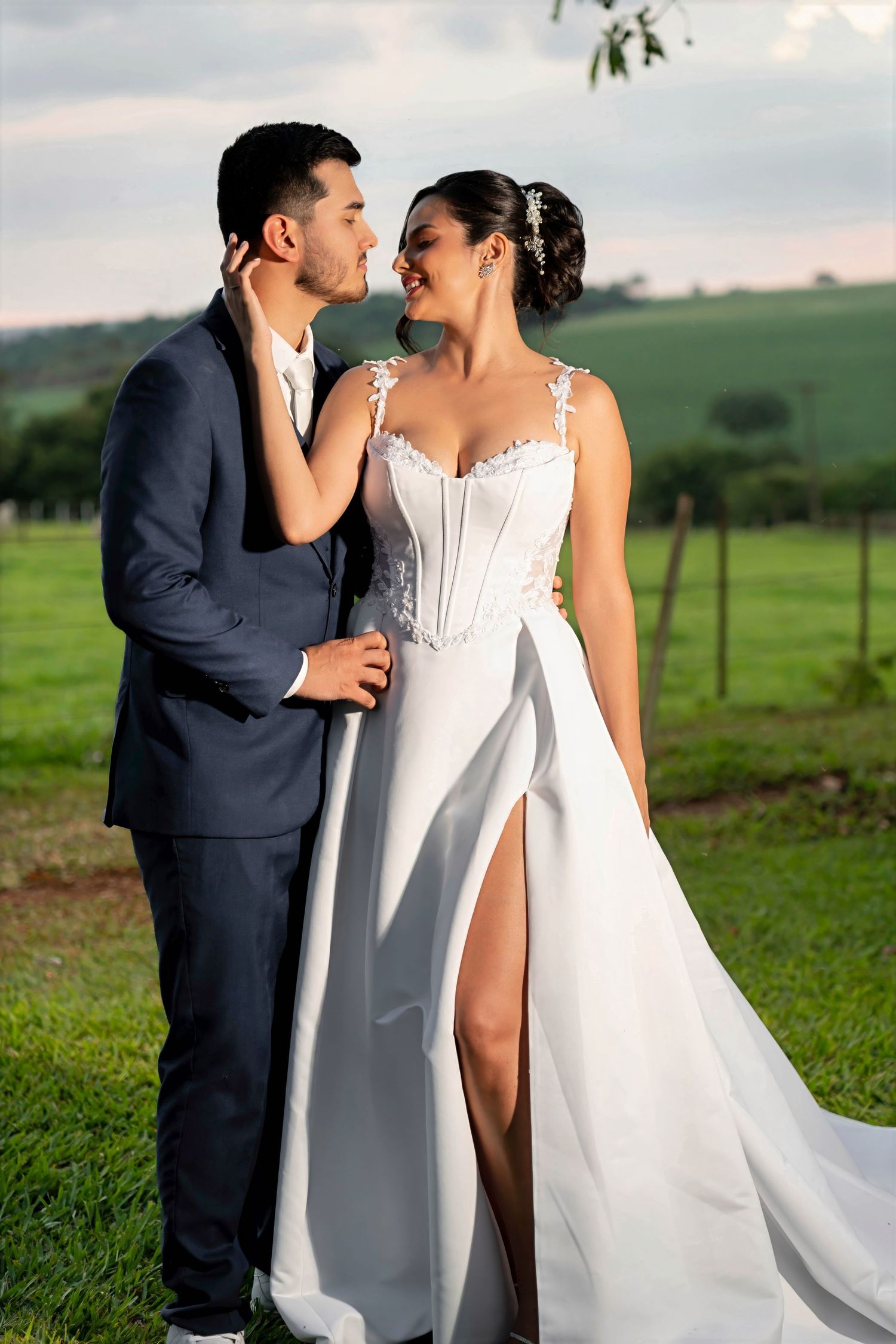 A bride in a white corset gown with a slit and groom in a navy suit share a romantic moment outdoors.