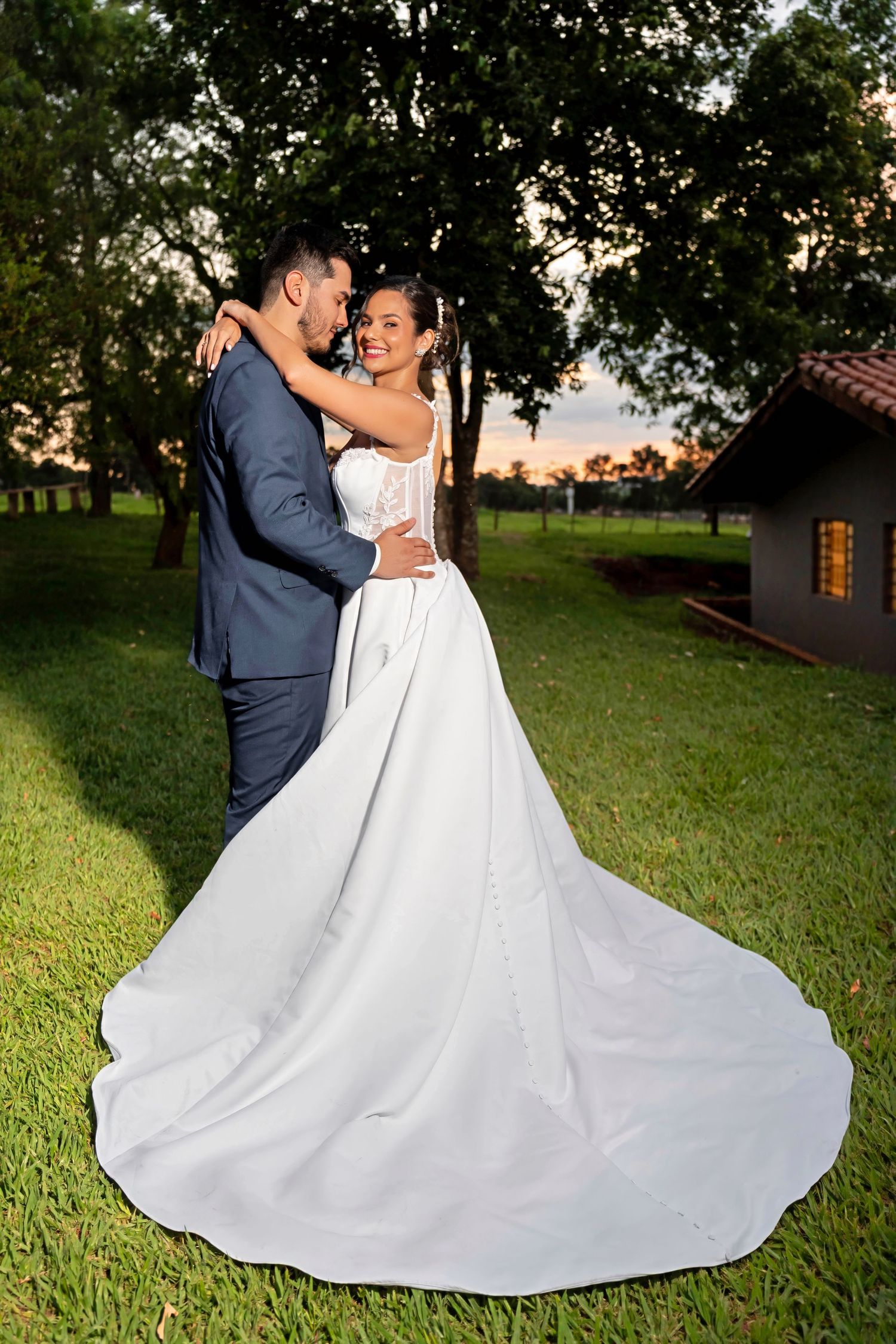 Bride in white gown with long train embraces groom in blue suit outdoors at sunset near trees.