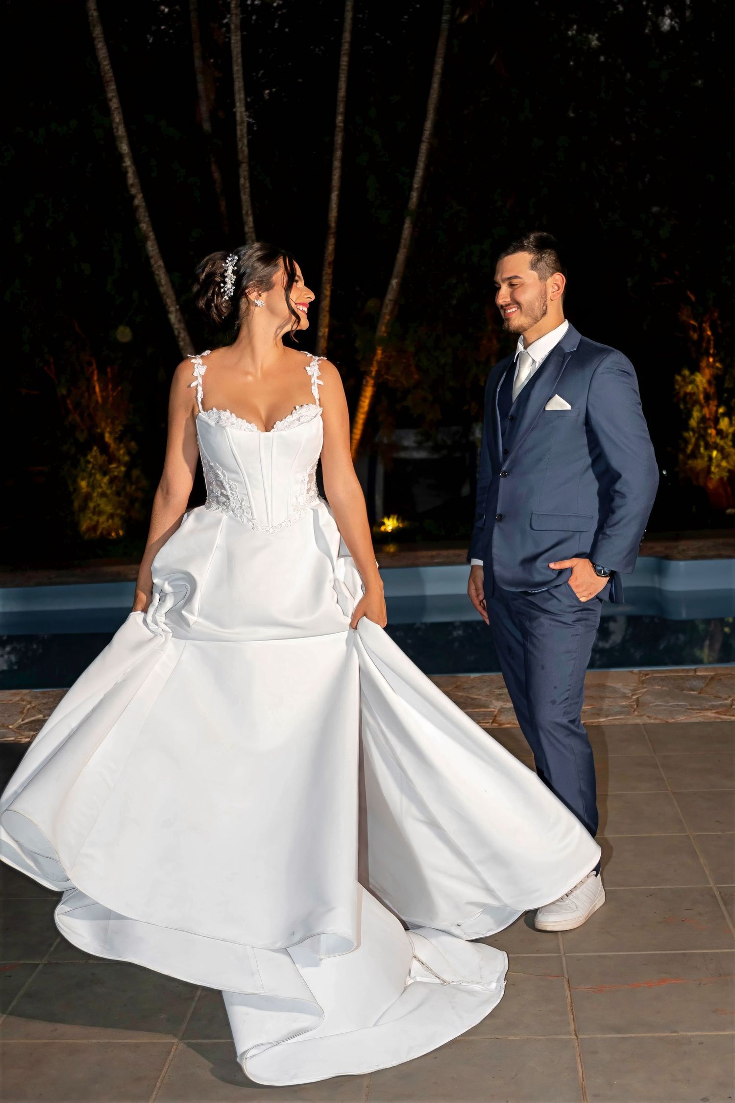 Bride in white gown twirling beside groom in navy suit near pool at night wedding celebration.
