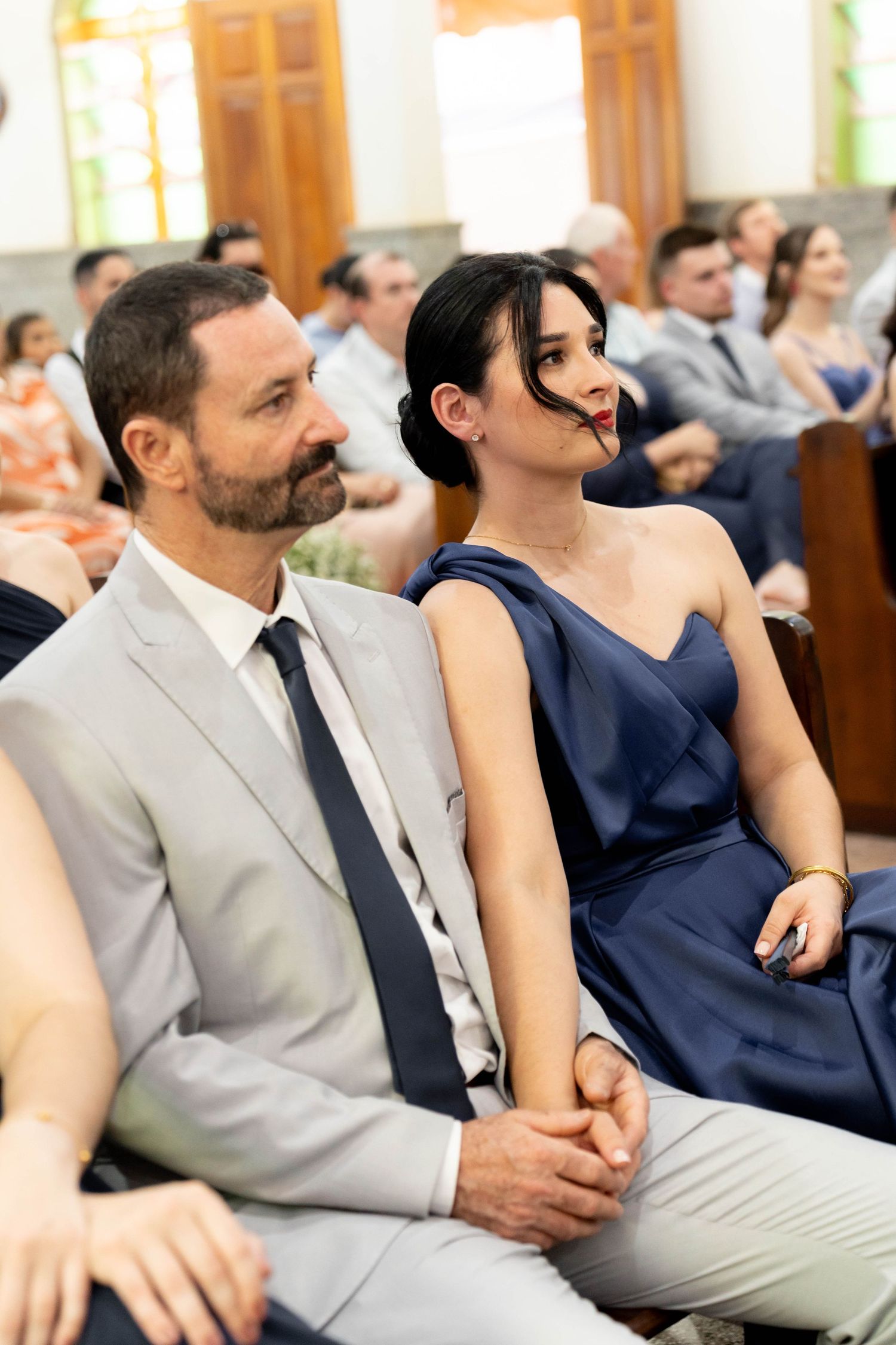 Couple in formal attire sitting together at a wedding ceremony, man in gray suit, woman in navy blue dress.