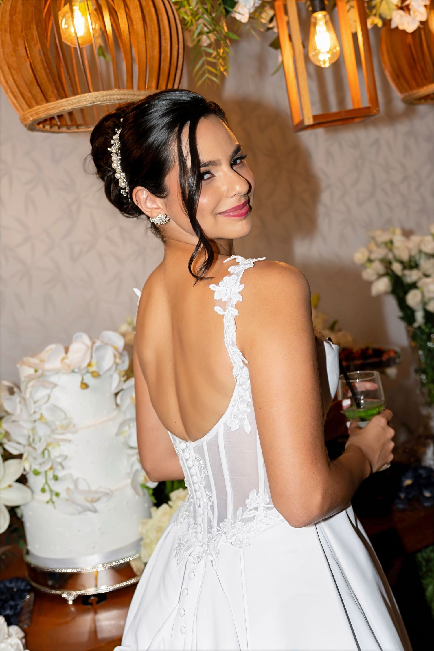 Bride in elegant backless white lace wedding gown smiling over shoulder near tiered cake holding champagne glass.