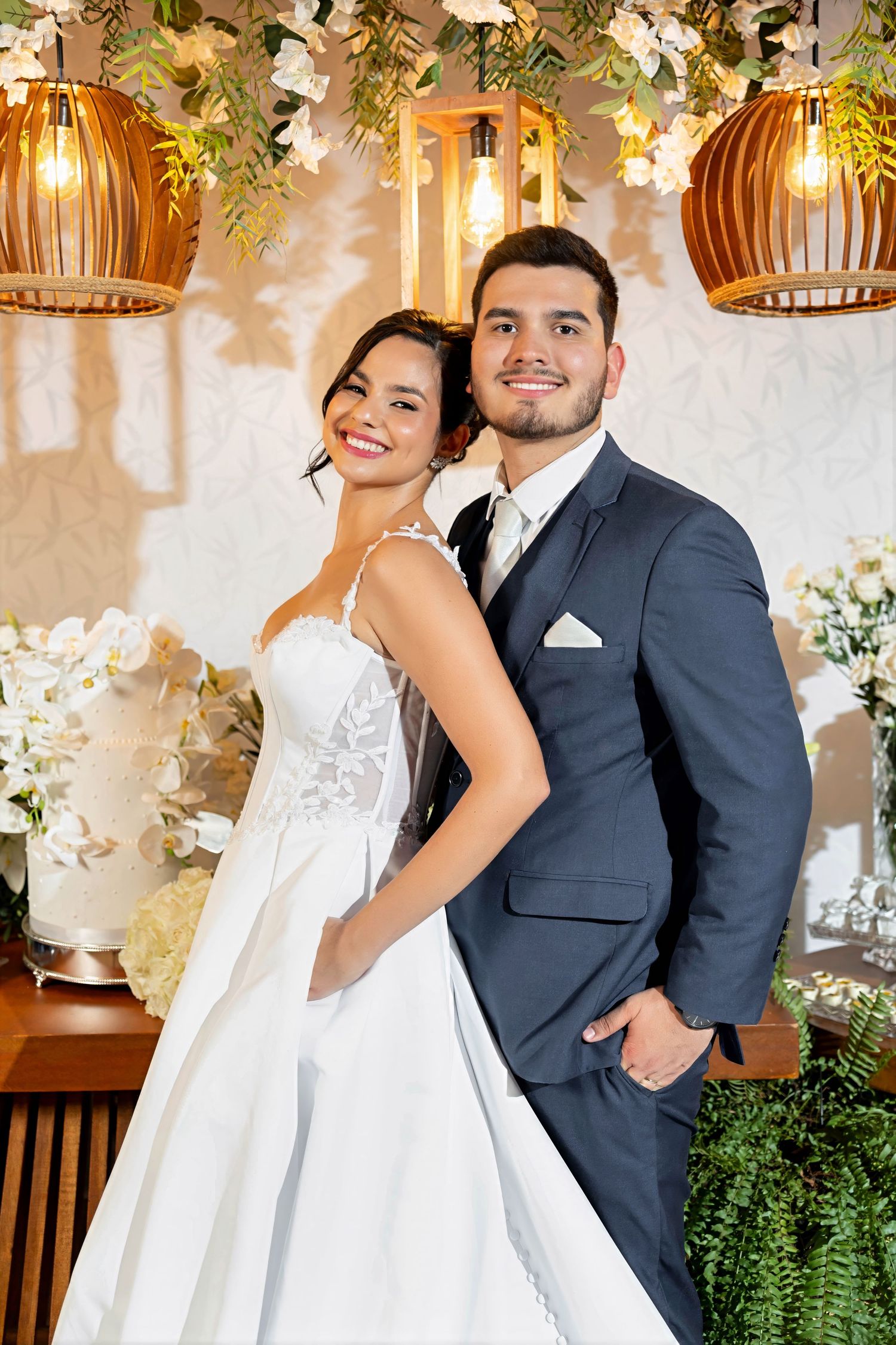 Smiling bride in white gown and groom in navy suit pose together at elegant wedding reception with floral decor.