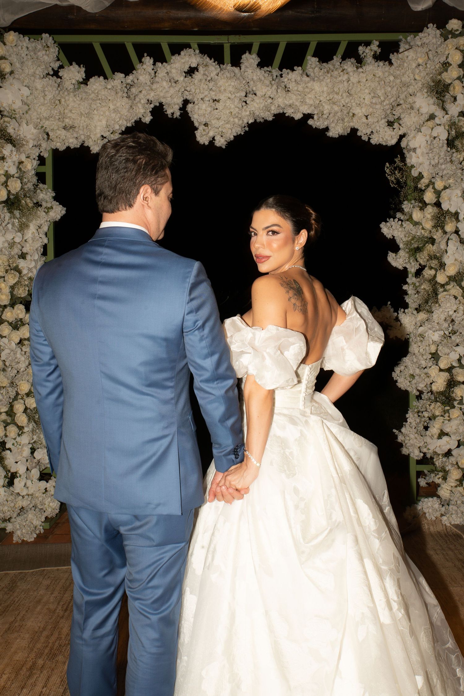 A bride in a white off-shoulder ballgown and groom in blue suit hold hands under a white floral arch at night.