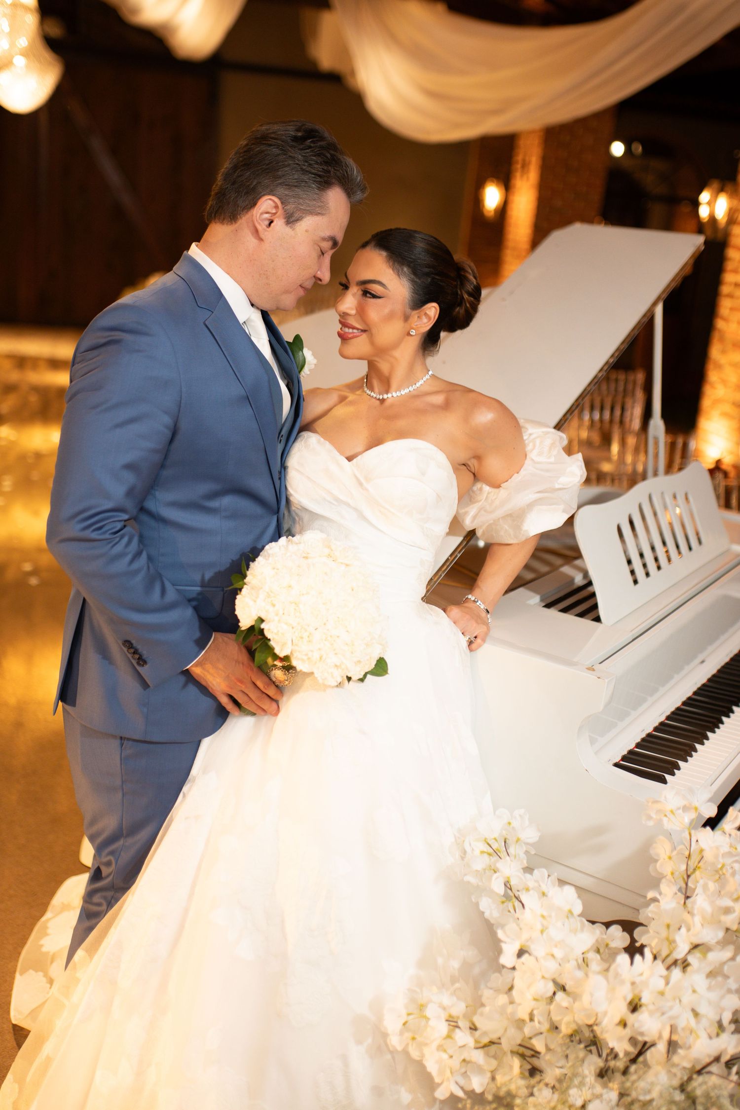 Bride in white ballgown holds bouquet beside groom in blue suit near white piano at elegant wedding reception.