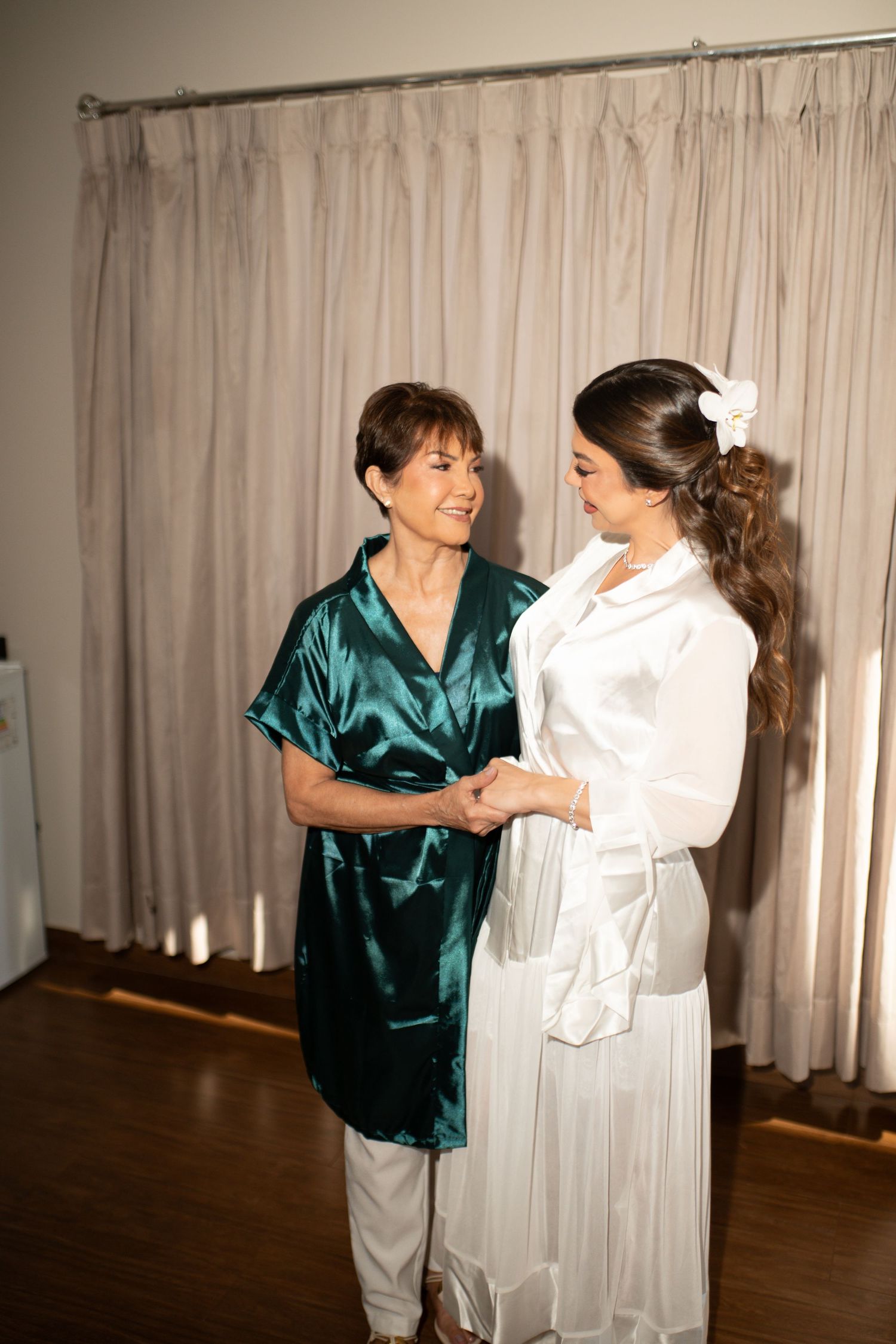 Two women in satin robes hold hands and smile, sharing a tender moment before a wedding ceremony.
