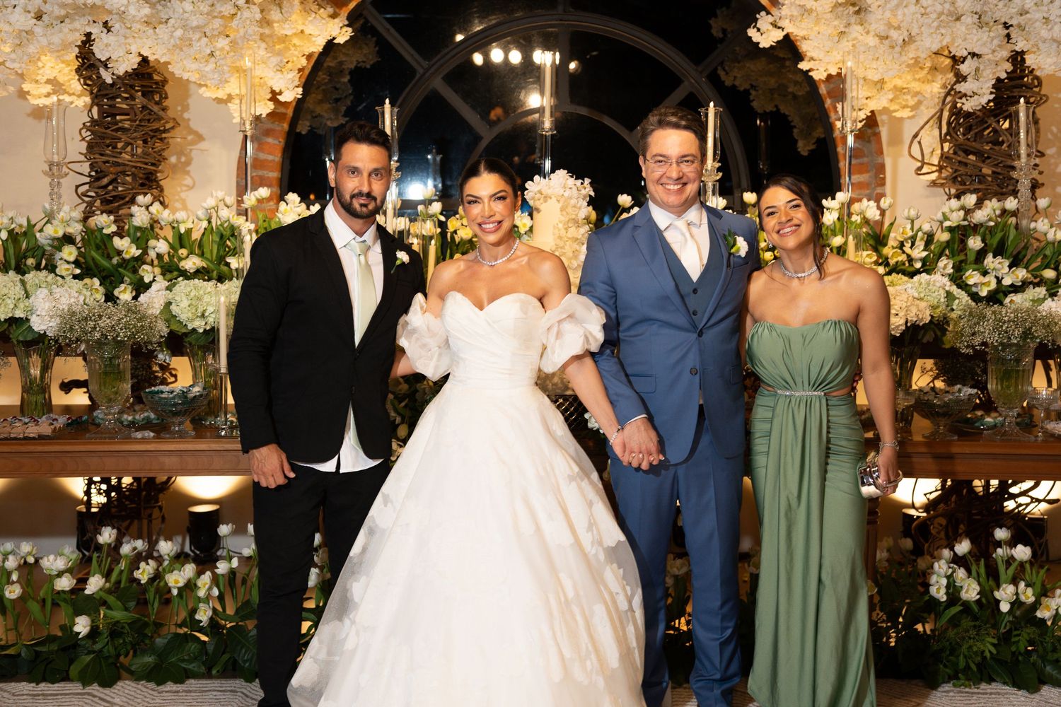 Elegant wedding photo with bride in white gown, groom in tuxedo, and guests in blue suit and green dress amid floral decor.