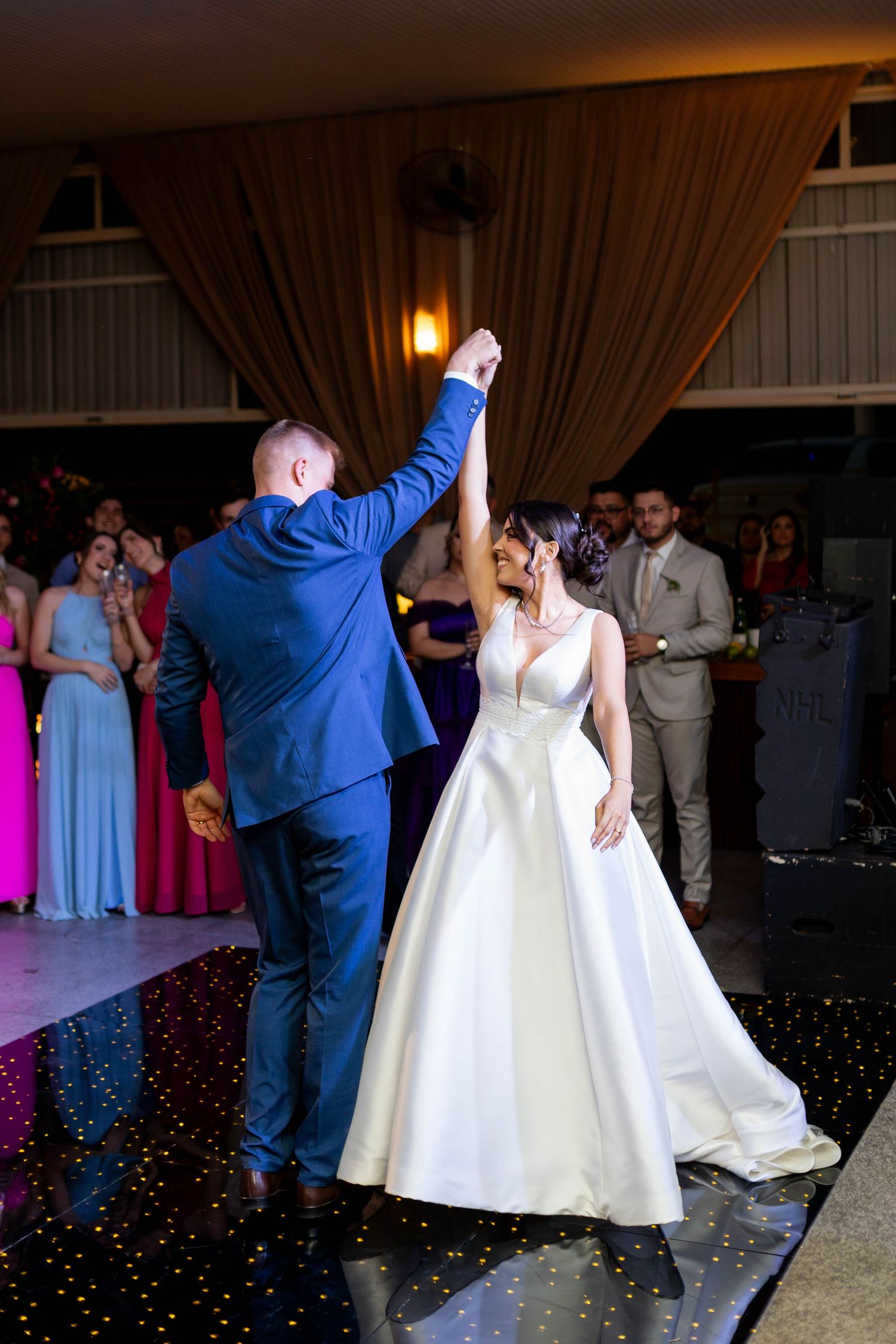 Bride and groom share their first dance at wedding reception, groom in blue suit spinning bride in white gown.