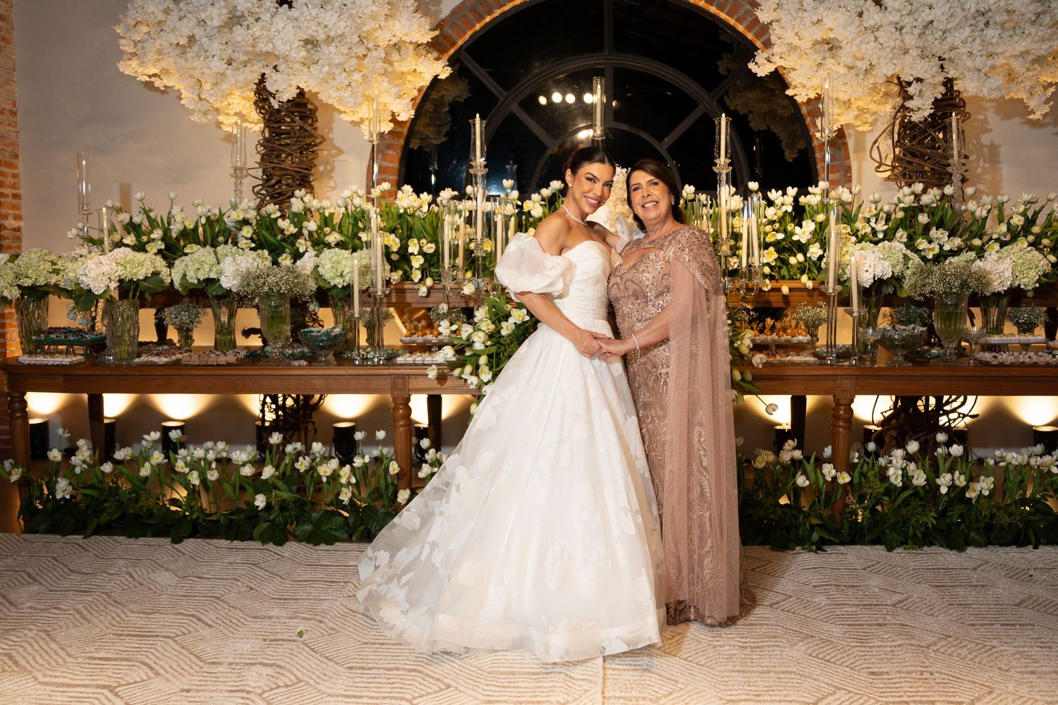Bride in white gown poses with elegant woman in gold dress before floral wedding backdrop with candles.