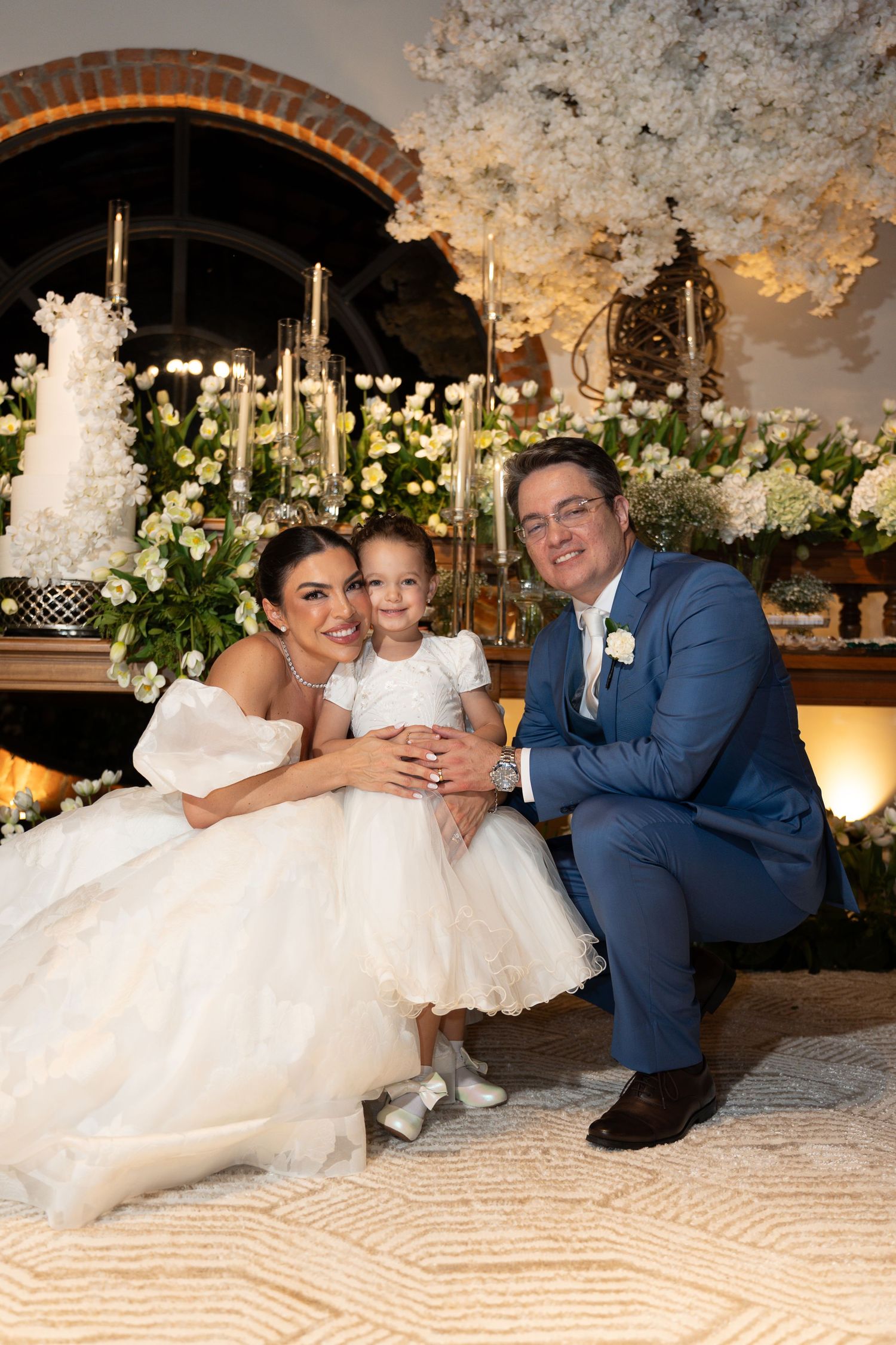 Bride and groom crouching with young flower girl in white dress at elegant wedding reception with floral backdrop.