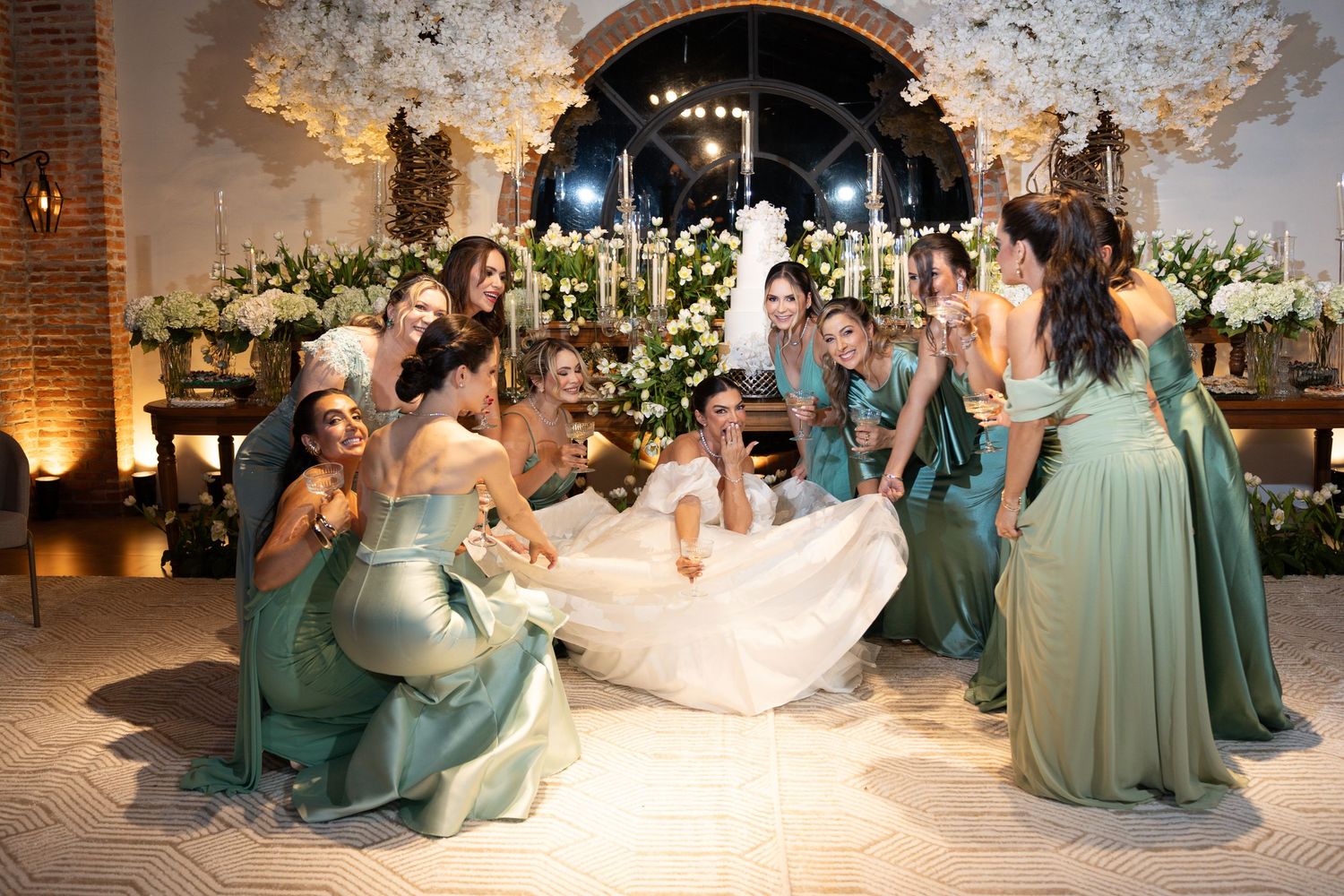 Bride in white gown surrounded by smiling bridesmaids in teal dresses at elegant floral wedding reception.