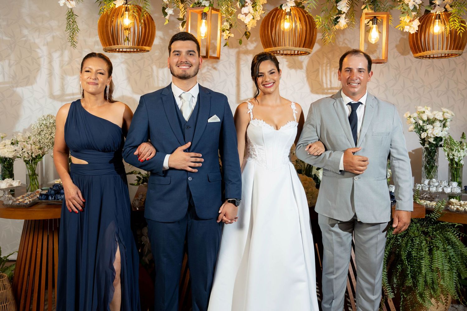 Wedding party of four posing elegantly, bride in white gown, groom in navy suit, flanked by family in formal attire.