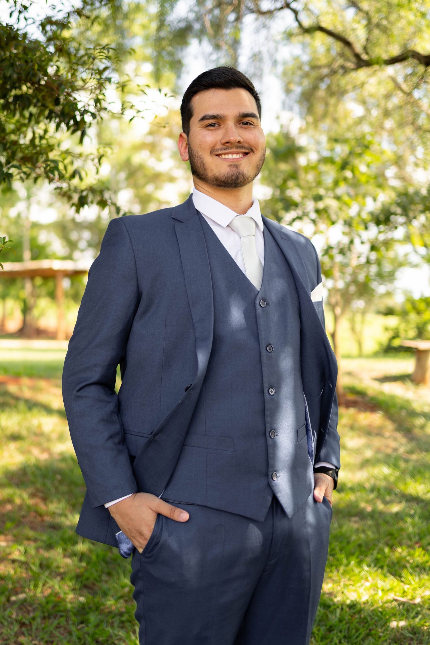 Young man in navy blue three-piece suit smiling outdoors in a garden setting.