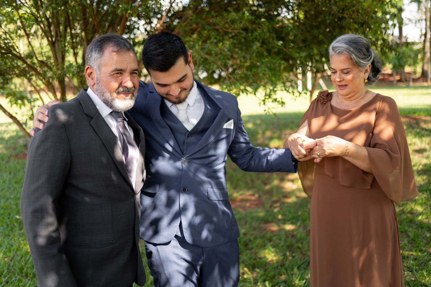 Groom in blue suit with elderly parents examining wedding ring outdoors in green park setting.