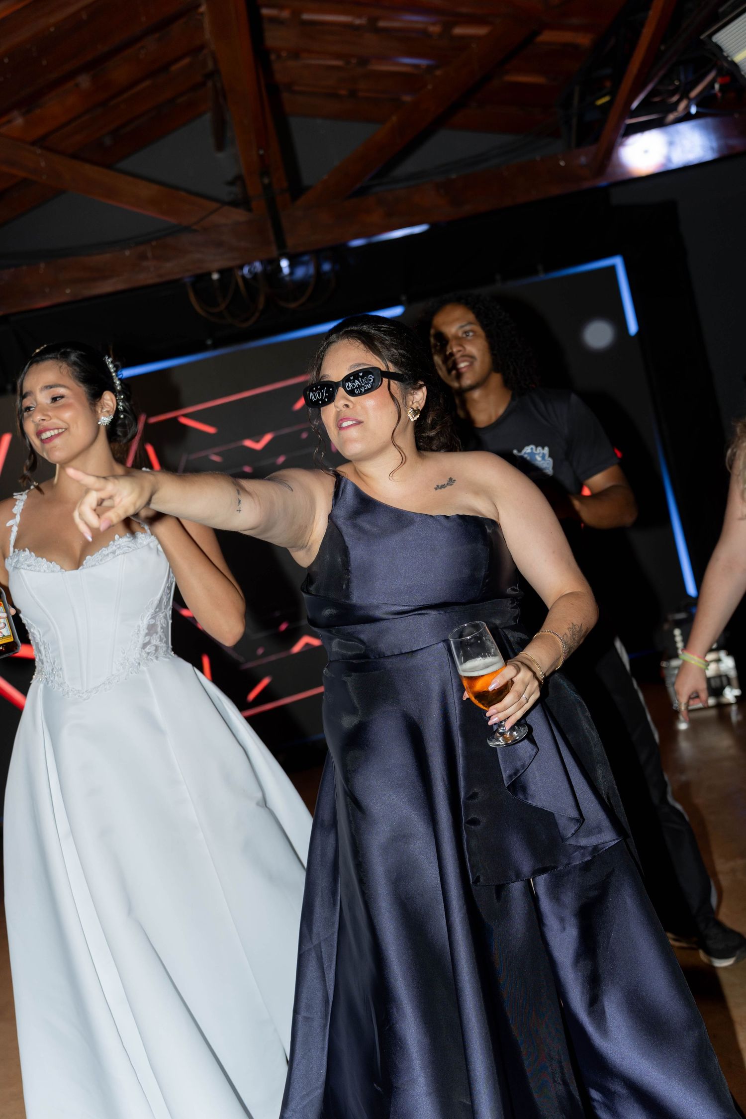 Bride in white gown dancing with guest in navy dress wearing sunglasses at wedding reception party.