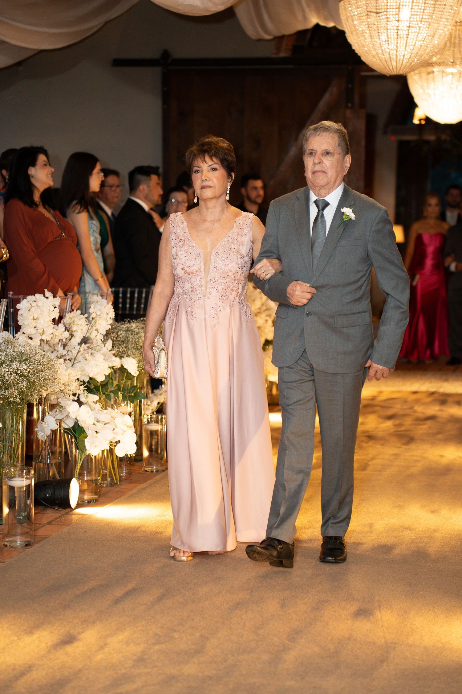 Elegant couple walking down wedding aisle, woman in blush gown, man in gray suit, floral arrangements and guests in background.