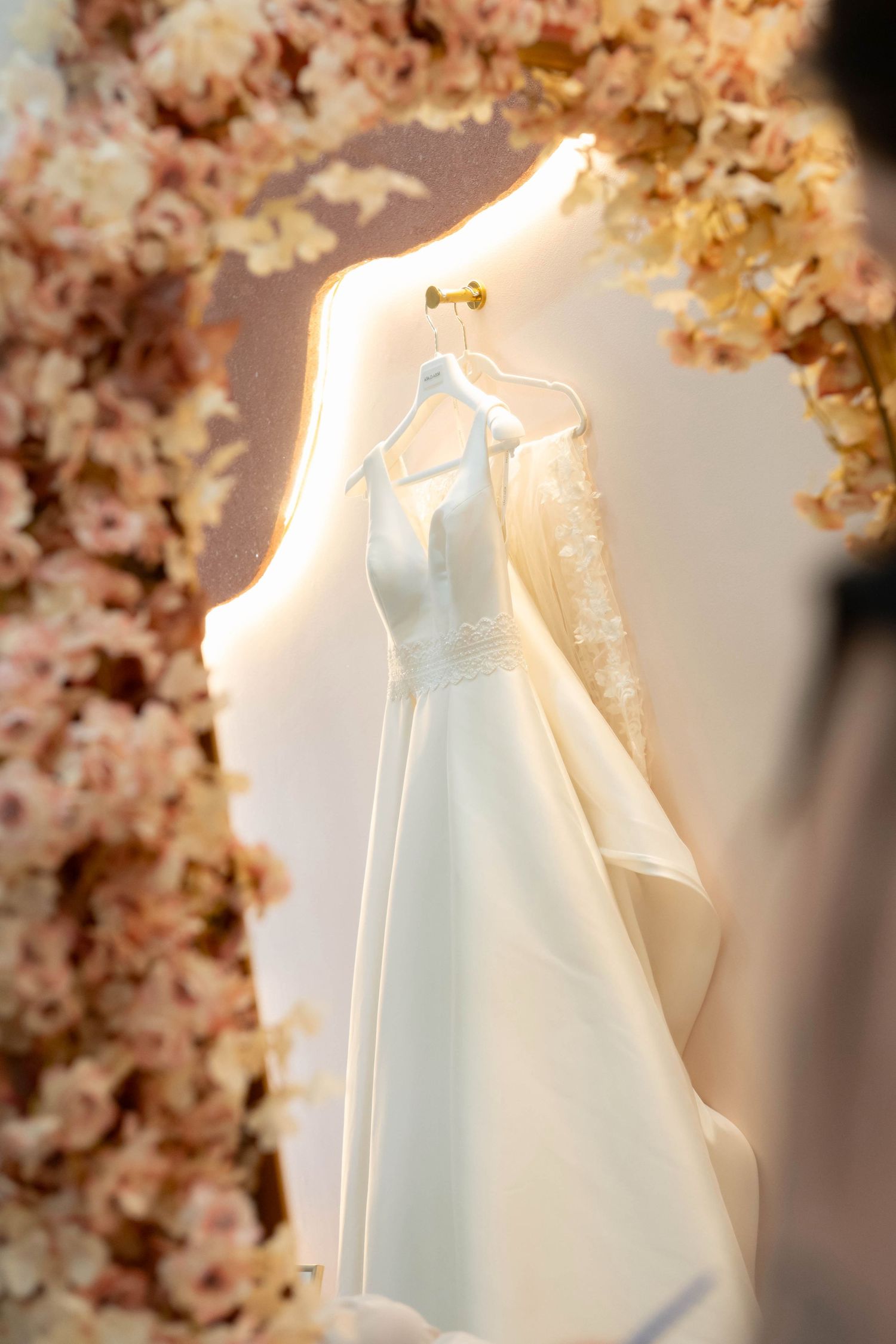 Elegant white wedding dress on a hanger displayed in a floral arch with soft pink blooms and warm lighting.