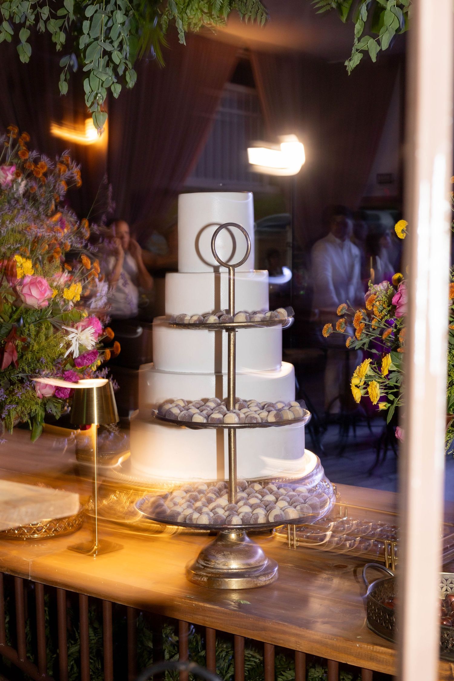 Elegant four-tier white wedding cake on silver stand with interlocked rings topper, surrounded by colorful floral arrangements.